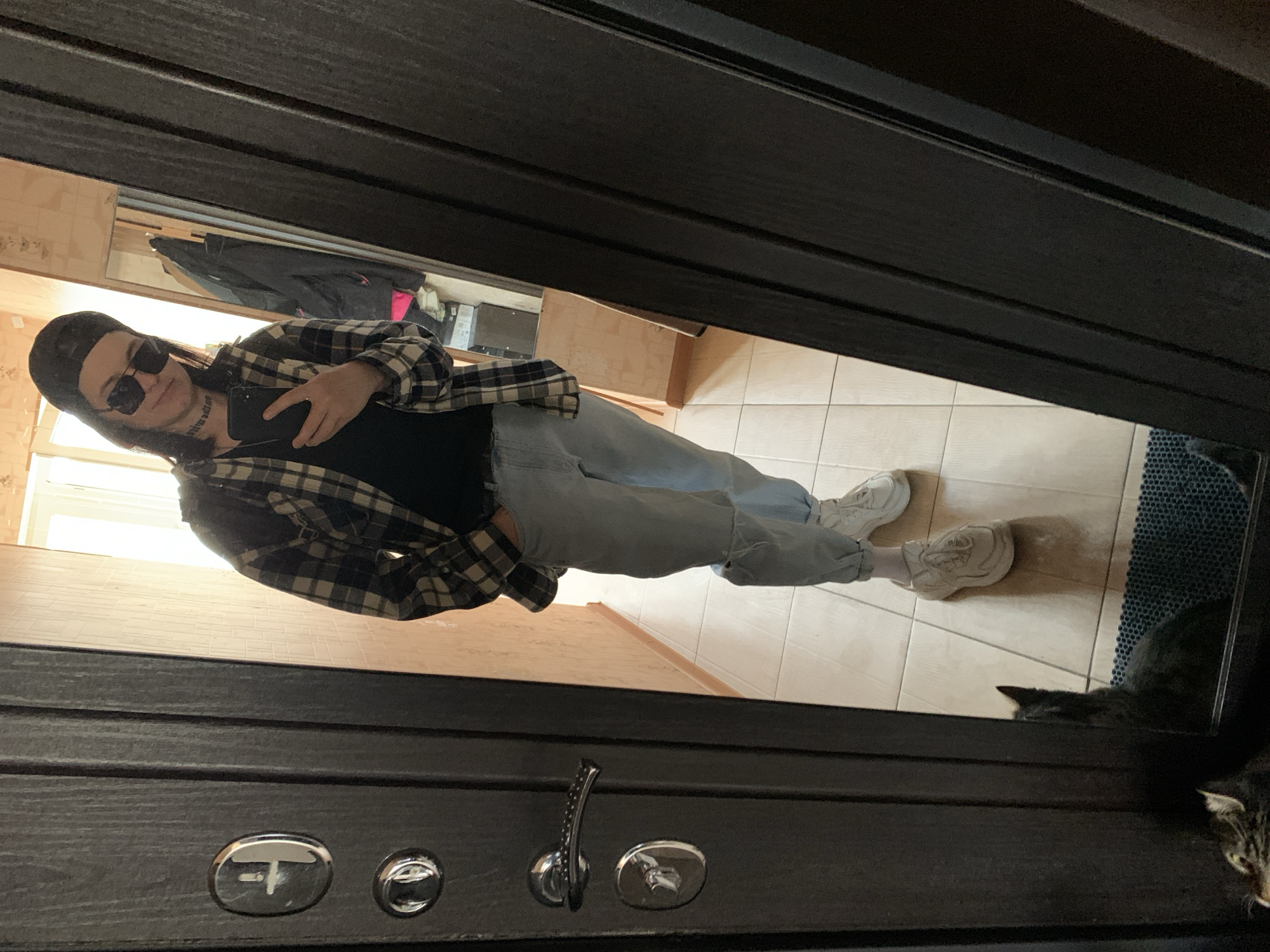 This image is a selfie captured in a reflective dark-colored door, rotated 90 degrees counter-clockwise. The primary subject is a person with long dark hair, wearing a dark cap, black sunglasses, a black and white plaid shirt over a black top, light blue jeans, and white chunky sneakers. They are holding a dark smartphone to take the photo. Unreadable text is tattooed on their neck.
The reflection in the door shows an indoor setting with light-colored tiled flooring, a window providing natural light, and some background objects including what appears to be a clothes rack with jackets and possibly a wooden box.
Two cats are visible in the foreground. A black cat is seen in the reflection on the tiled floor, near a dark blue mat. Closer to the actual camera, a tabby cat is partially visible, looking upwards. The dark door itself features several metallic components, including a handle, locks, and a possible peephole. The scene is well-lit, indicating it is daytime.