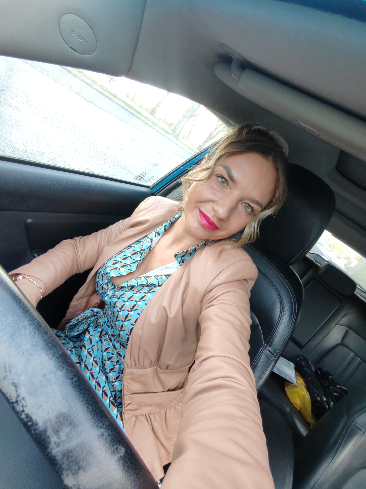 A woman sits in the driver's seat of a car. She wears a blue patterned dress, a tan leather jacket, and bright pink lipstick. Her hair is pulled back in a bun. The interior of the car is black leather. The airbag symbol is visible on the car's ceiling. A plastic bag is on the back seat. The scene appears to be in a car, and given the limited visual information, determining the exact location in Petropavlovsk-Kamchatsky is not possible.