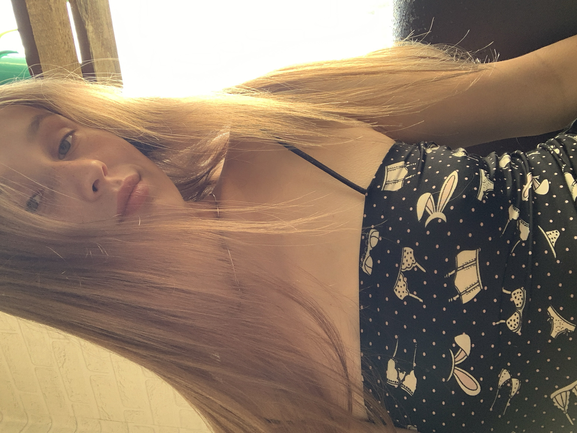 This is a close-up, sideways portrait of a young woman with long, blonde hair. She is lying down, with her head tilted back and to the side, exposing her face and upper chest. Her hair is spread out around her head and shoulders, appearing slightly windswept. She is wearing a dark strapless top with a pattern of small pink polka dots and white line drawings of undergarments, including bras and panties, and a pair of bunny ears. Her expression is relaxed and calm, with her eyes partially closed and a soft smile on her lips. The background is out of focus, but appears to be outdoors, with a hint of sunlight suggesting daytime. There are some wooden elements and a brick-like texture visible. The overall mood is casual and peaceful.