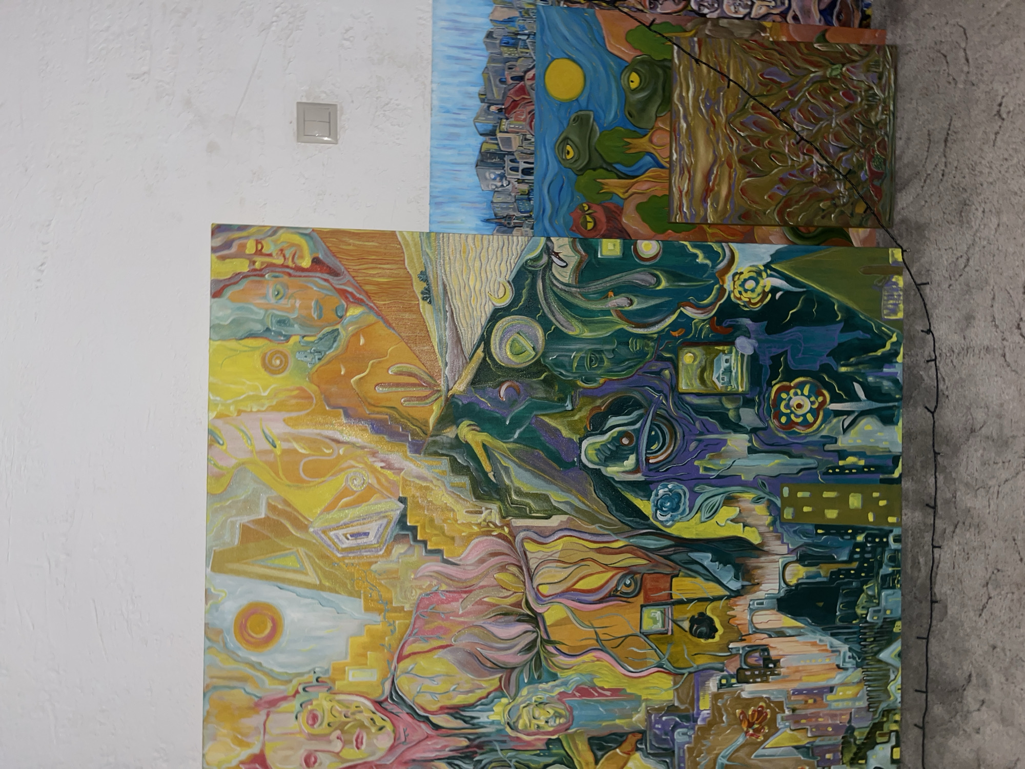 Several abstract paintings are displayed against a white and gray wall, with a light switch visible in the top left corner. The largest painting features a blend of yellow, orange, pink, green, and purple hues. A smaller painting above depicts a cityscape beside a river, with a yellow sun. A third painting, only partially visible, shows shades of brown, green, and purple. Thin black cords hang down the right side of the paintings. The location cannot be determined.
