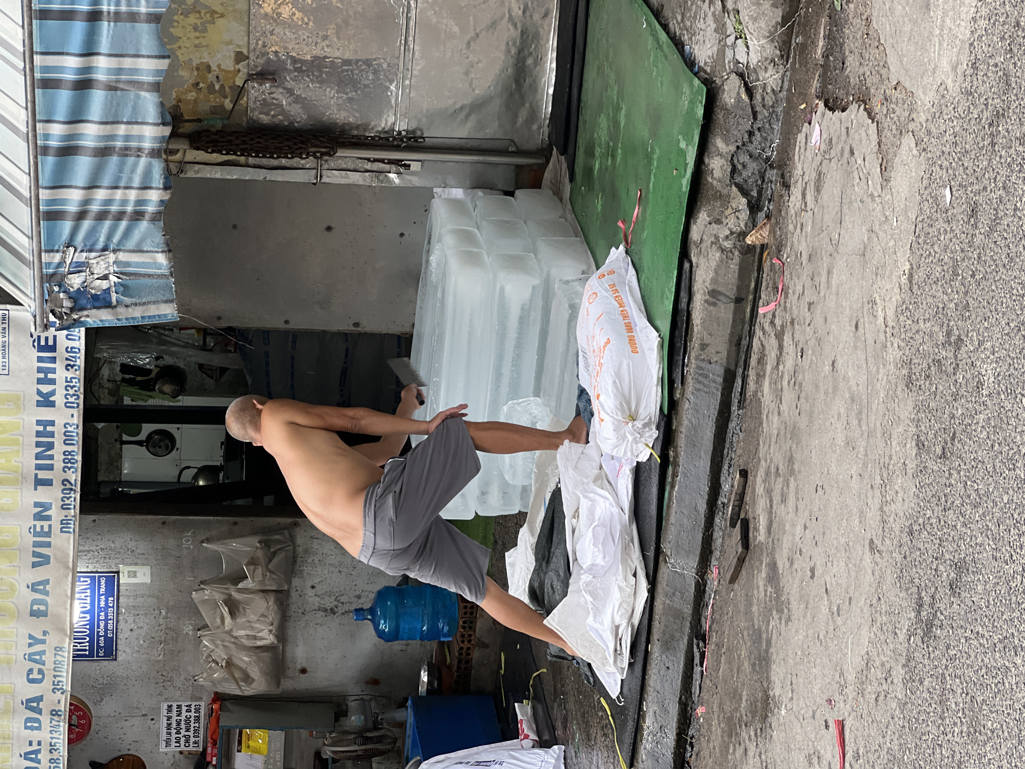 A man, shirtless and wearing gray shorts, is cutting large blocks of ice with a serrated knife. He is standing on a wet, paved surface next to a stack of ice blocks and a large blue water jug. There are several white plastic bags on the ground, some filled with ice. The scene appears to be on a street in Nha Trang, Vietnam, indicated by signage in Vietnamese that reads "TRUONG GIANG" and "ĐC: 60A ĐÔNG ĐA - NHA TRANG" which translates to "Address: 60A Dong Da - Nha Trang." The background shows a building with a blue and white striped awning and various items hanging from the ceiling. The ground is wet, suggesting recent rain or melting ice.