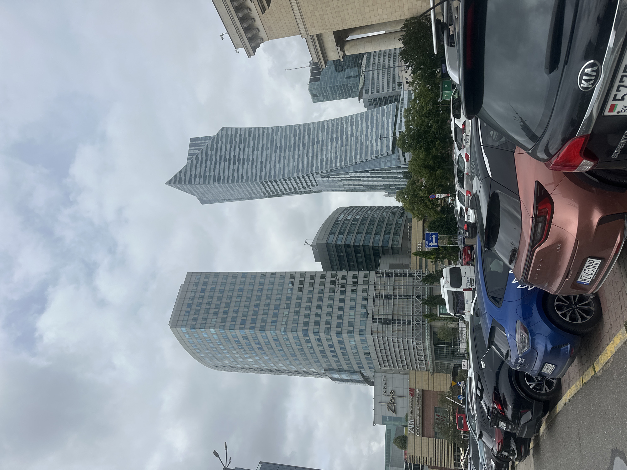 The image depicts a view of the central business district of Warsaw, Poland, characterized by modern skyscrapers and a parking lot filled with cars. In the background, several tall buildings dominate the skyline, including the distinctive warped skyscraper known as the Warsaw Hub, and another, more traditionally rectangular skyscraper to its left. The building with the most prominent architectural feature is a tall, glass-covered skyscraper with a striking, twisted design. To the left of this, a very tall, flat-sided skyscraper with a grid-like pattern of windows is visible, identified by signage for the Złote Tarasy shopping mall. Several other buildings of varying heights and architectural styles are also present. In the foreground, a street-level view shows a parking lot filled with a variety of cars, mostly SUVs and hatchbacks, parked parallel to the street. The license plates are partially visible on some vehicles, with one plate clearly showing Polish characters. The sky is overcast, indicating a cloudy day. The overall scene suggests an urban environment with a mix of commercial and retail spaces, bustling with activity.