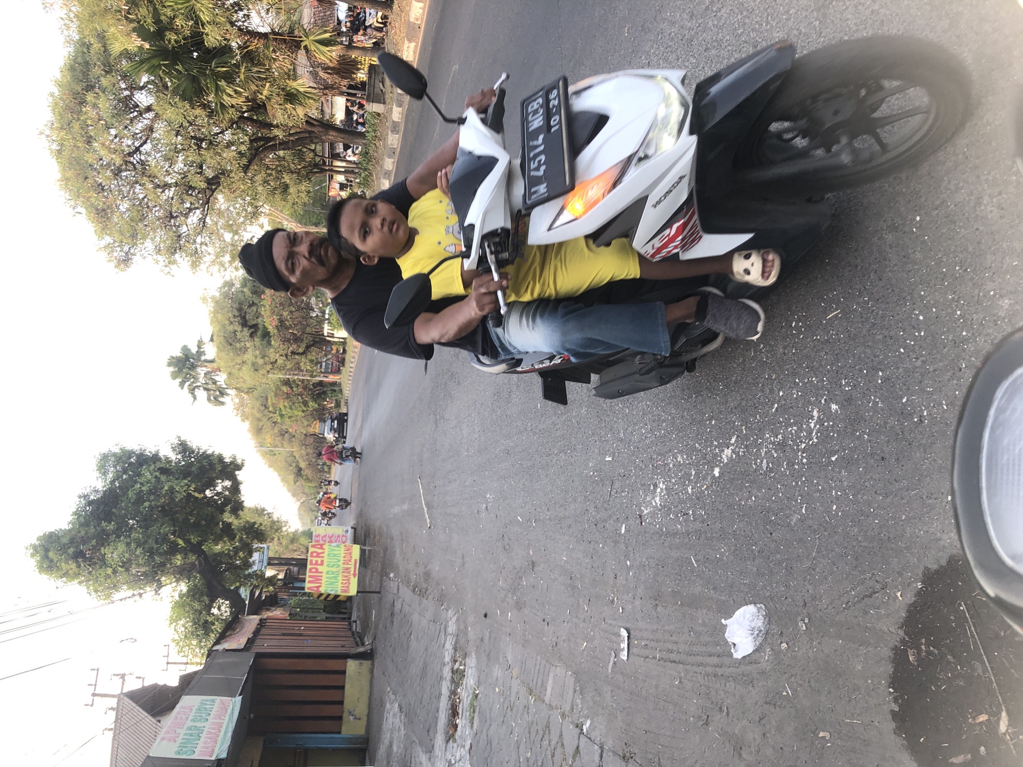 In Ngaban, Indonesia, a man wearing a black beanie and a black t-shirt rides a white Honda motorcycle, giving a ride to a young boy in a yellow shirt. The man is driving the motorcycle on a paved road, with trees and buildings lining the sides. The boy is seated in front of the man, with his arms around the man's waist. The motorcycle has a license plate that reads "H 4514 NGB". The scene appears to be during the day, with sunlight filtering through the trees. There are other vehicles and people in the background, suggesting that this is a public street. The overall impression is one of a common mode of transportation and everyday life in Indonesia.