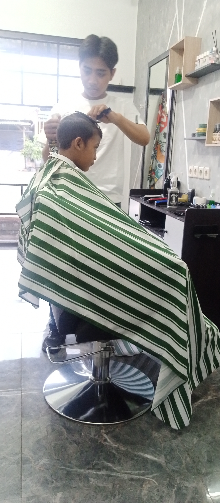 The image depicts a barbershop scene in Lajuk, Indonesia. A young boy is seated in a barber chair, draped with a green and white striped cape, receiving a haircut from a barber. The barber, wearing a white t-shirt, is in the process of cutting the boy's hair with clippers and a comb. The barbershop appears to be well-lit, with a large window in the background showing an outdoor scene. Shelving units with various barber supplies are visible on the wall, along with a mirror. The floor is tiled with a marble-like pattern.