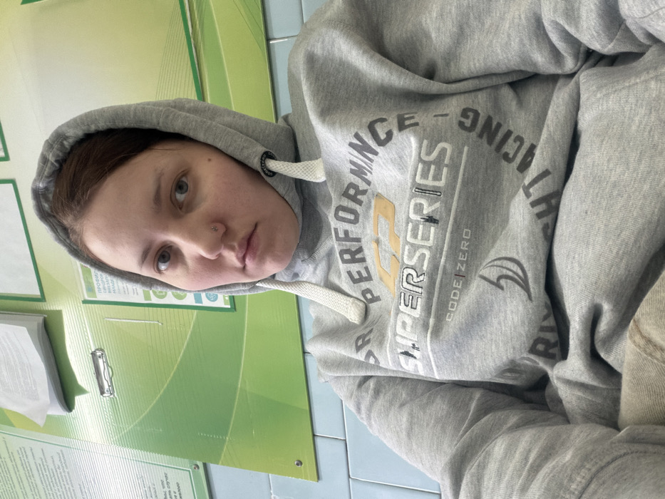A person with fair skin and a nose piercing wears a gray hooded sweatshirt. The sweatshirt features the words "PERFORMANCE" and "SUPER SERIES" in a graphic design. The person looks directly at the camera. The background includes a green wall with posters or signs. The person is likely indoors.