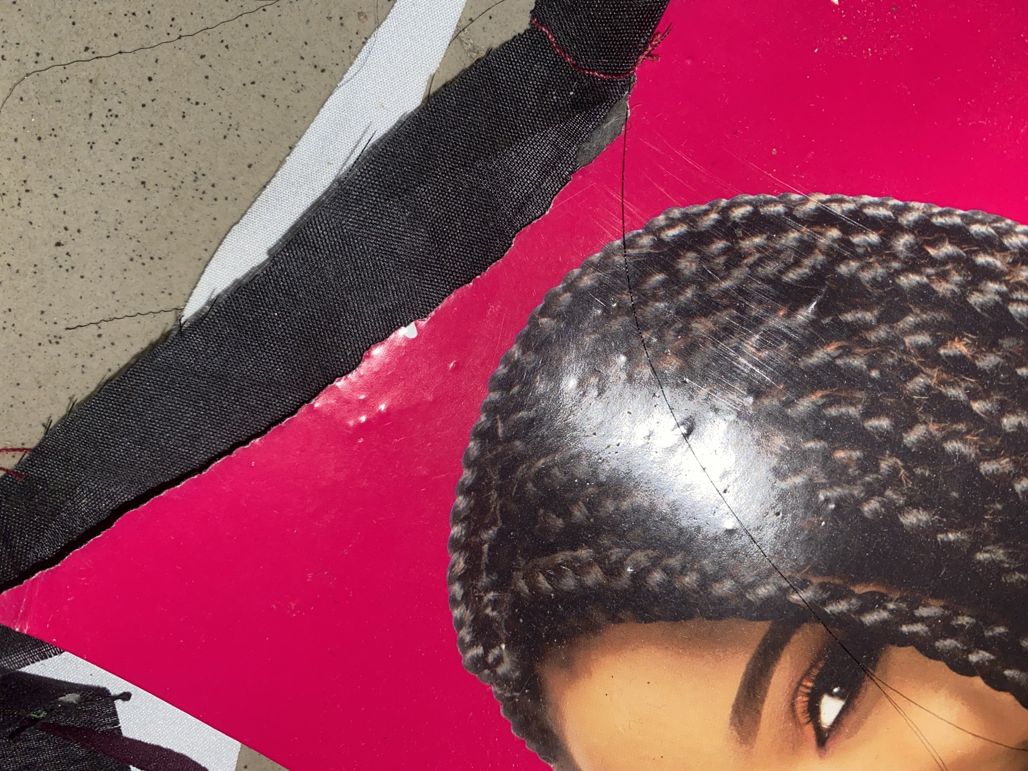 The image displays a close-up of a portion of a bright pink surface, possibly a poster or magazine, featuring the eye and braided hair of a person. The person's skin tone is dark. The pink surface appears to be torn and has scratches and scuff marks. Overlapping the pink surface are pieces of black and white fabric, and what appears to be a rough, speckled concrete or stone surface. There are no immediate indications of Jauro Yinu, Nigeria.