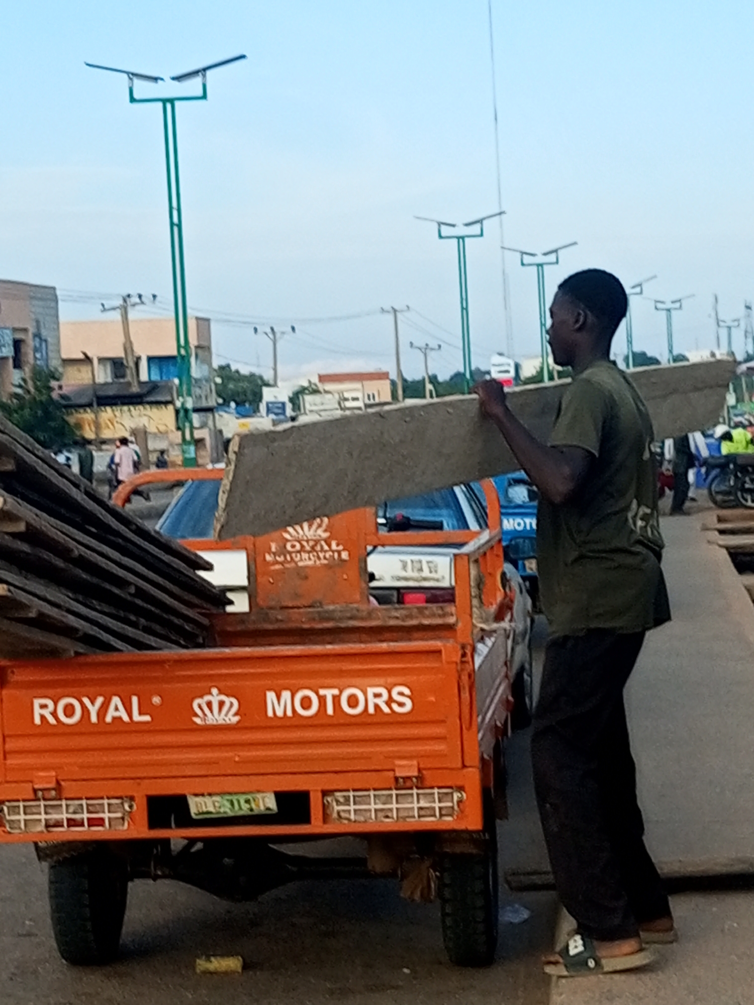 In Gombe, Nigeria, a young Black man wearing a dark green t-shirt and dark pants is carrying a long, flat, light gray object, possibly a plank of wood or concrete, on his right shoulder. He is walking on a paved surface next to an orange, three-wheeled cargo truck with "ROYAL MOTORS" written on the back. The truck is loaded with stacked wooden planks. In the background, several streetlights with solar panels are visible, along with buildings, power lines, and other vehicles, including motorcycles. The scene appears to be outdoors in a semi-urban environment, possibly a street or a market area.