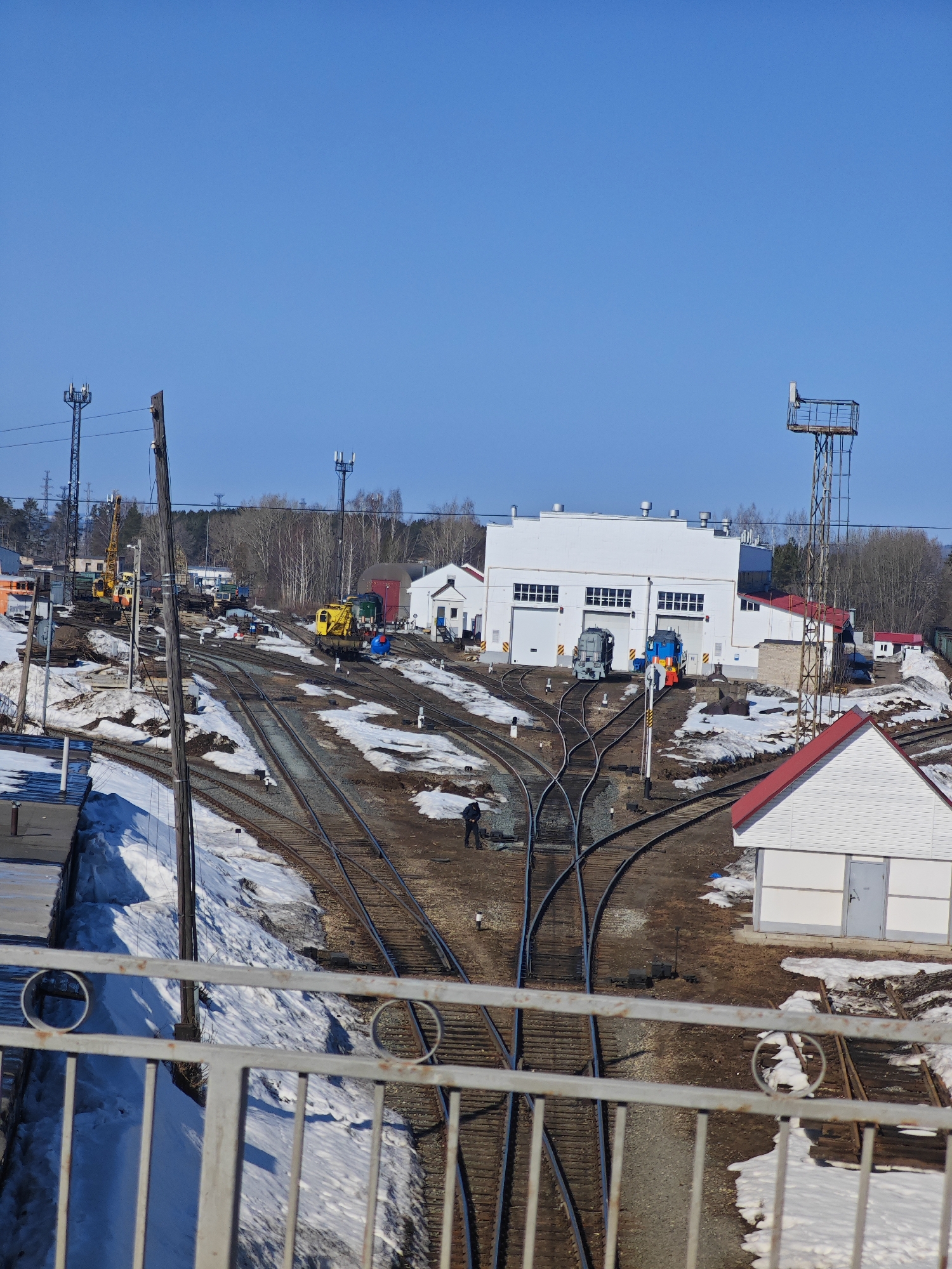 The image depicts a railway yard in Chaikovsky, Russia, in early spring, with remnants of snow on the ground. Multiple sets of railway tracks converge and diverge, leading towards a large, white industrial building with several garage-like doors. Various pieces of railway maintenance equipment are scattered throughout the yard, including a yellow crane and several locomotives. A person is walking along the tracks near a smaller, white building with a red roof. Overhead, a clear blue sky contrasts with the scene below. A metal guardrail is visible in the foreground, indicating the photo was taken from an elevated perspective, possibly a bridge.