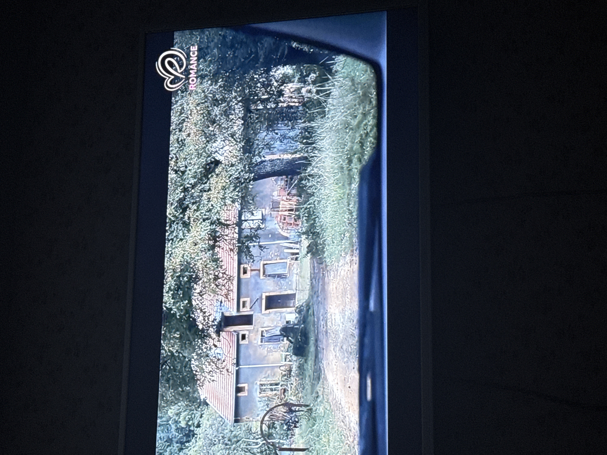 A TV screen displays a scene of a rustic building viewed from an obscured path. The "Romance" channel logo is visible in the upper left corner. The building, likely an old house or farmhouse, has a faded blue or gray exterior with a red tiled roof. There are multiple windows and doors visible, some seemingly boarded up or in disrepair. Dense foliage, including trees and overgrown vegetation, surrounds the building and obscures the path leading to it. The path is lightly overgrown. The scene appears to be filmed from a moving vehicle or handheld device, creating a blurred effect along the edge of the frame. Given the presence of the "Romance" channel logo and the rustic setting, it is likely from a romantic movie or TV show. There is not enough information to determine its location in the city of Mytishchi, Russia.
