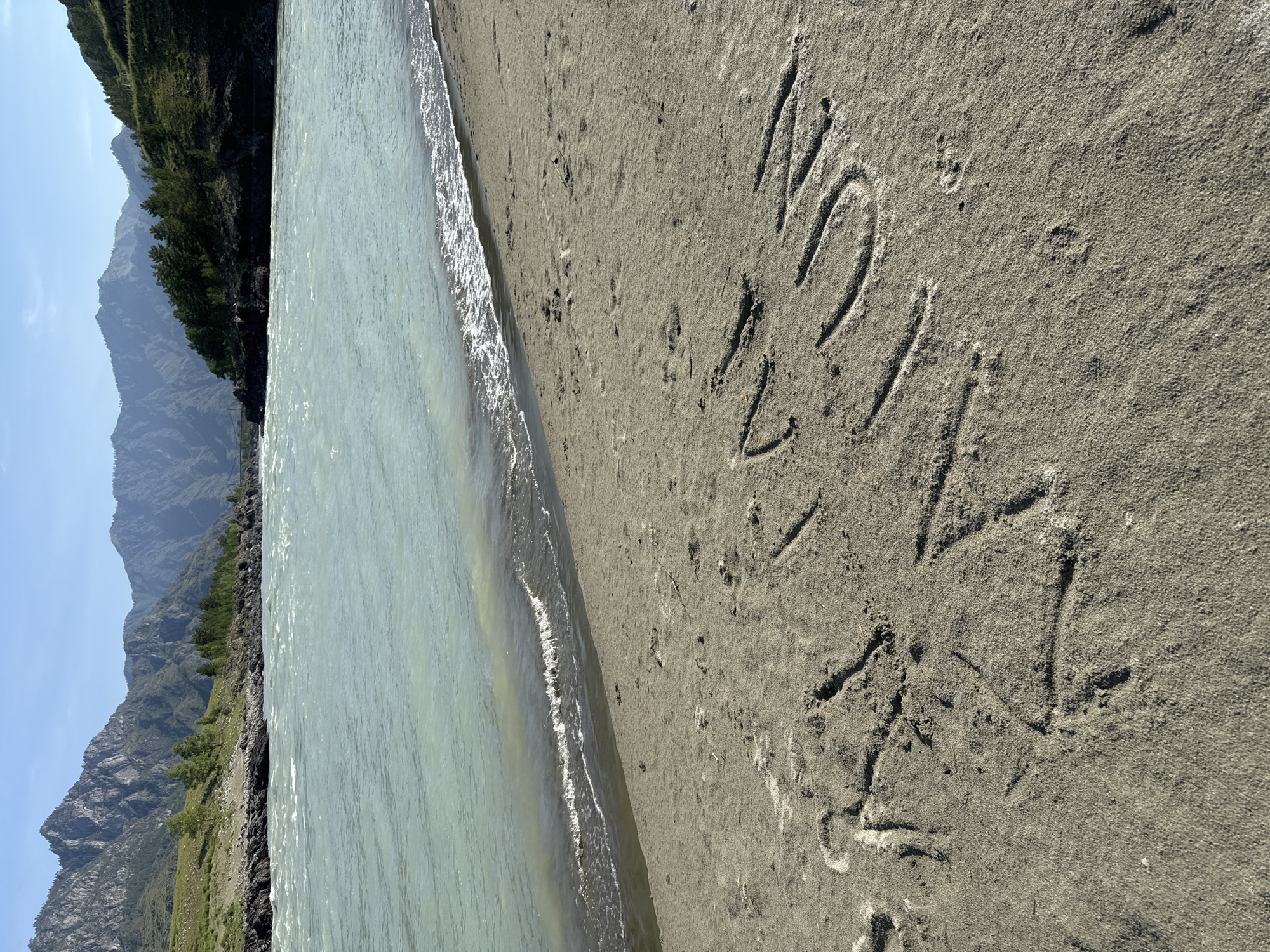 In Edigan, Russia, on a sandy bank of a wide, turquoise river, text is inscribed in the sand. The text, written in large, uneven letters, appears to read "MAYA". Mountains covered in trees form the background, with a bright, clear sky above. The river exhibits ripples and some wave activity near the shore. The sand shows footprints and some debris. The image is captured from a low angle, focusing on the sand and the inscribed text in the foreground, with the river and mountains in the background.