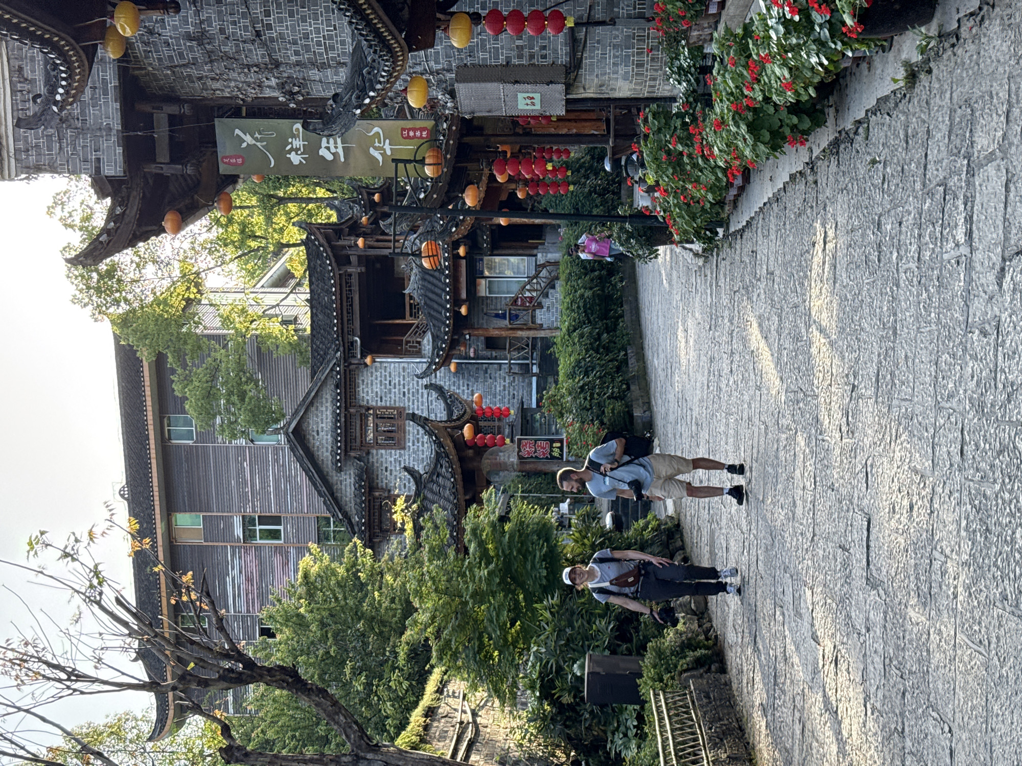 The image shows a narrow, cobblestone street in Furong, China. Traditional Chinese architecture lines both sides of the street, with multi-tiered buildings, ornate roofs, and red lanterns. Two men are walking along the street, seemingly tourists, as one carries a camera. Red flowers hang above the street on one side. The location is likely within the ancient town section of Furong.