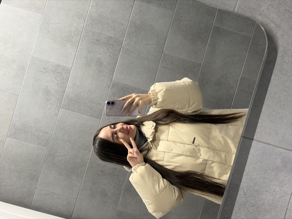 A single female individual is depicted in a mirror selfie within an indoor setting. The subject has long, dark hair and is wearing a light beige puffer jacket over a grey turtleneck. Her left hand is raised, forming a V-sign. Her right hand holds a light purple smartphone with a decorative case, positioned to capture her reflection. The individual exhibits a slight smile or smirk with partially closed eyes. The background consists of a wall covered in large, rectangular, dark grey tiles arranged horizontally. A tiled floor is partially visible in the reflection at the bottom of the mirror. The image contains no visual information to determine a specific location within the city of Novosaratovka, Russia.