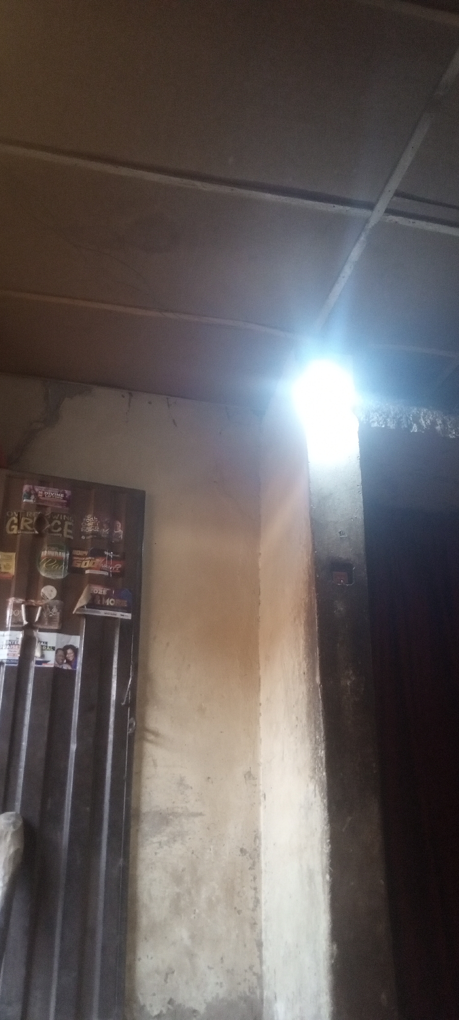 This image captures an interior scene, likely a room or passage in Sabon Tasha, Nigeria, characterized by stark lighting and simple construction. No people are visible.
To the left, a corrugated metal door or gate stands, adorned with several papers, posters, and stickers. Visible text includes "DIVINE LIVING" (partially seen on a top-left poster), "OVERFLOWING GRACE" (larger text below the previous), and "ABUNDANT CURRY" (on a green label). Other smaller, less legible texts can be seen on various flyers, including one that appears to say "HOME" and another with "International..."
The wall to the left of a prominent central concrete pillar is light-colored, possibly unpainted concrete or plaster, showing signs of wear and slight discoloration. A very bright, intense light source, emanating from the upper right corner, creates significant glare and casts strong illumination on the upper part of the wall, the ceiling, and the right side of the central pillar. This pillar forms a sharp corner, leading into a dark, recessed area or doorway on the far right.
The ceiling above is a light brown-grey color, featuring visible beams or structural elements. The strong light suggests either a powerful artificial light or direct sunlight streaming into the space. The overall impression is of a functional, perhaps utilitarian, indoor environment.