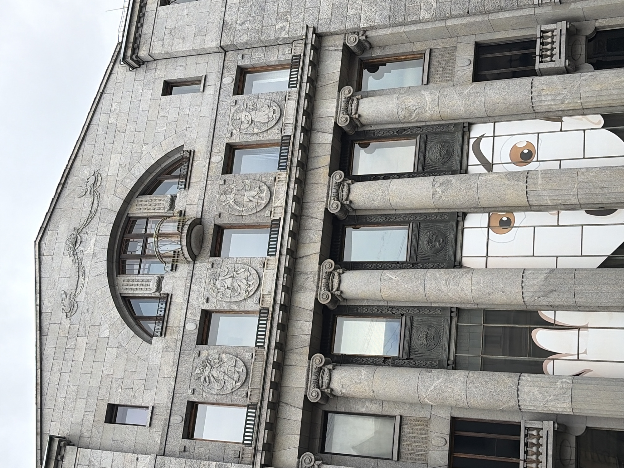 The image shows a close-up view of the facade of a grand stone building in Saint Petersburg, Russia. The building features classical architectural elements such as large fluted columns with ornate capitals, arched windows, and decorative stone carvings. The facade is constructed from light-colored stone blocks, giving it a monolithic appearance.
In the upper part of the building, there is a large arched window with a small balcony projecting from it. Below this, several rectangular windows are visible, each adorned with circular stone reliefs depicting figures. The lower section of the building has more windows, some of which are covered by large, stylized graphics of eyes and a hand. These graphics appear to be part of a temporary advertisement or art installation, contrasting with the building's traditional architecture. The sky above is overcast, with a pale, diffused light. The overall impression is one of historical grandeur overlaid with a modern, playful element.