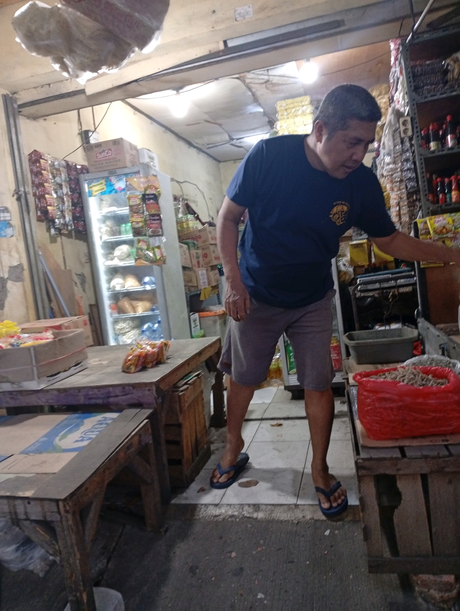 The image depicts a man standing in a small shop, likely a market stall, in North Jakarta, Indonesia. The man is wearing a dark blue t-shirt and grey shorts. He is reaching towards a large red bag filled with small items, possibly dried fish or snacks, which is placed on a wooden crate. In front of him is a wooden table with a box on it. To his left, a glass-fronted refrigerator is visible, stocked with various products. Shelves lined with goods are seen in the background. The environment appears to be an indoor market setting with concrete flooring. The lighting is artificial, suggesting an interior space.