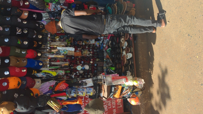 The image displays an outdoor market stall in Moshi, Tanzania, under bright daylight. The stall, filling the upper portion of the frame, is densely packed with merchandise. The top section showcases multiple rows of baseball caps and beanies, many displaying various logos and circular reflective stickers. Below these, a vertical display features numerous small items including keychains, watches, necklaces, and small bags, often against a red background. The bottom section of the stall consists of a multi-tiered rack holding numerous pairs of sandals and open-toed shoes. On the left side, a partial red sign with white text, reading "TUNASA LINE" and "NA KUREN LINE," is visible. An olive-green beanie hangs nearby.
An adult male, viewed from behind, stands facing the merchandise. He wears a dark short-sleeved t-shirt with a yellow stripe on the shoulder, dark grey jeans, and an orange knit cap. He is barefoot, standing on a reddish-brown, unpaved, and uneven ground surface composed of dirt and small stones. Shadows, indicative of strong sunlight, are cast onto the ground. A small, light-colored stone and a dark object lie on the ground near his feet. The man appears to be observing the items on display.