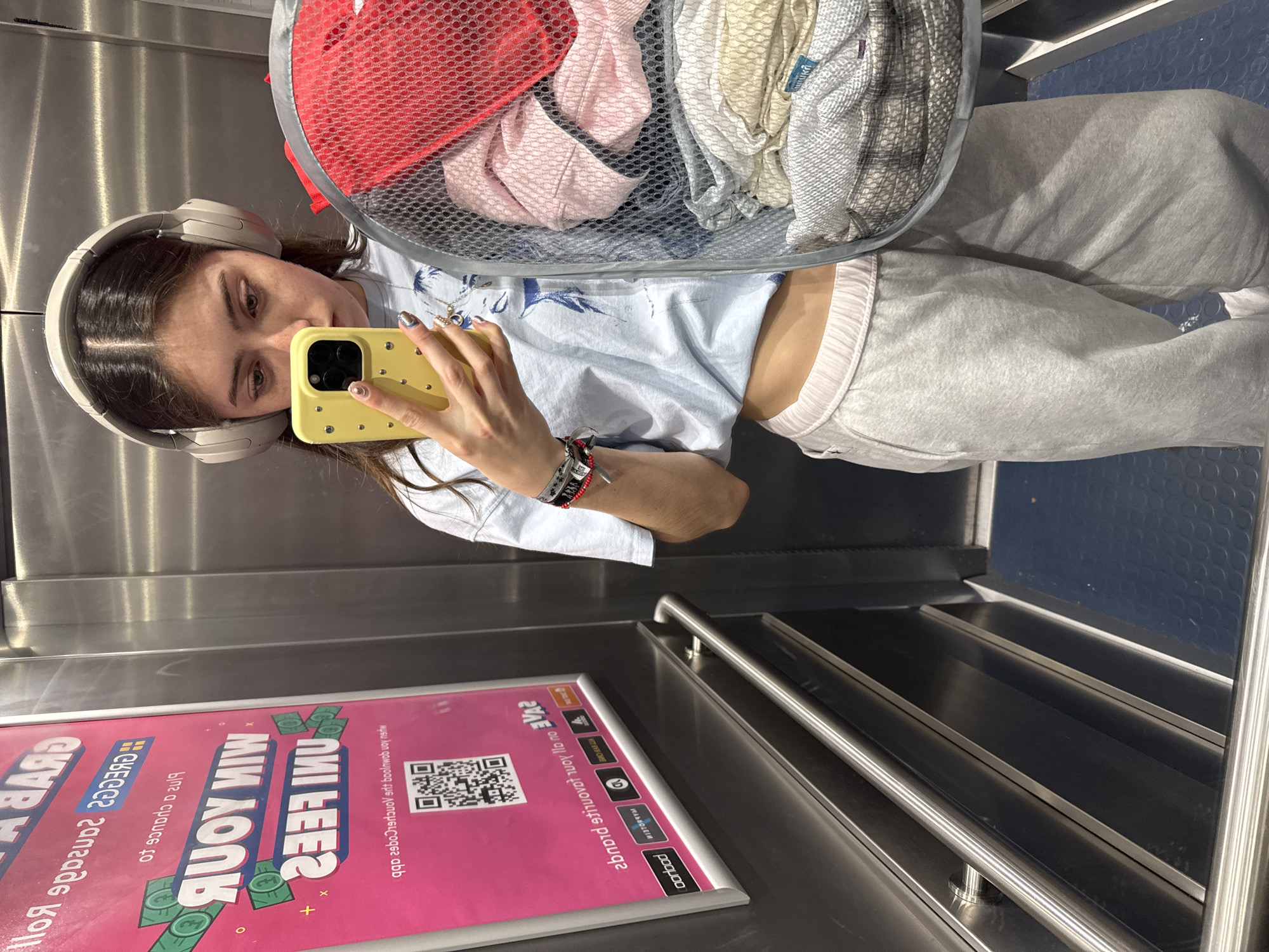 A woman is taking a selfie in an elevator with a yellow phone. She is wearing headphones and a light blue shirt with gray sweatpants. She is holding a laundry basket full of clothes. A promotional sign is visible on the elevator wall with an offer to "WIN YOUR FEES." The sign also contains a QR code. The elevator is located in a shopping centre in the city of Hull, United Kingdom.