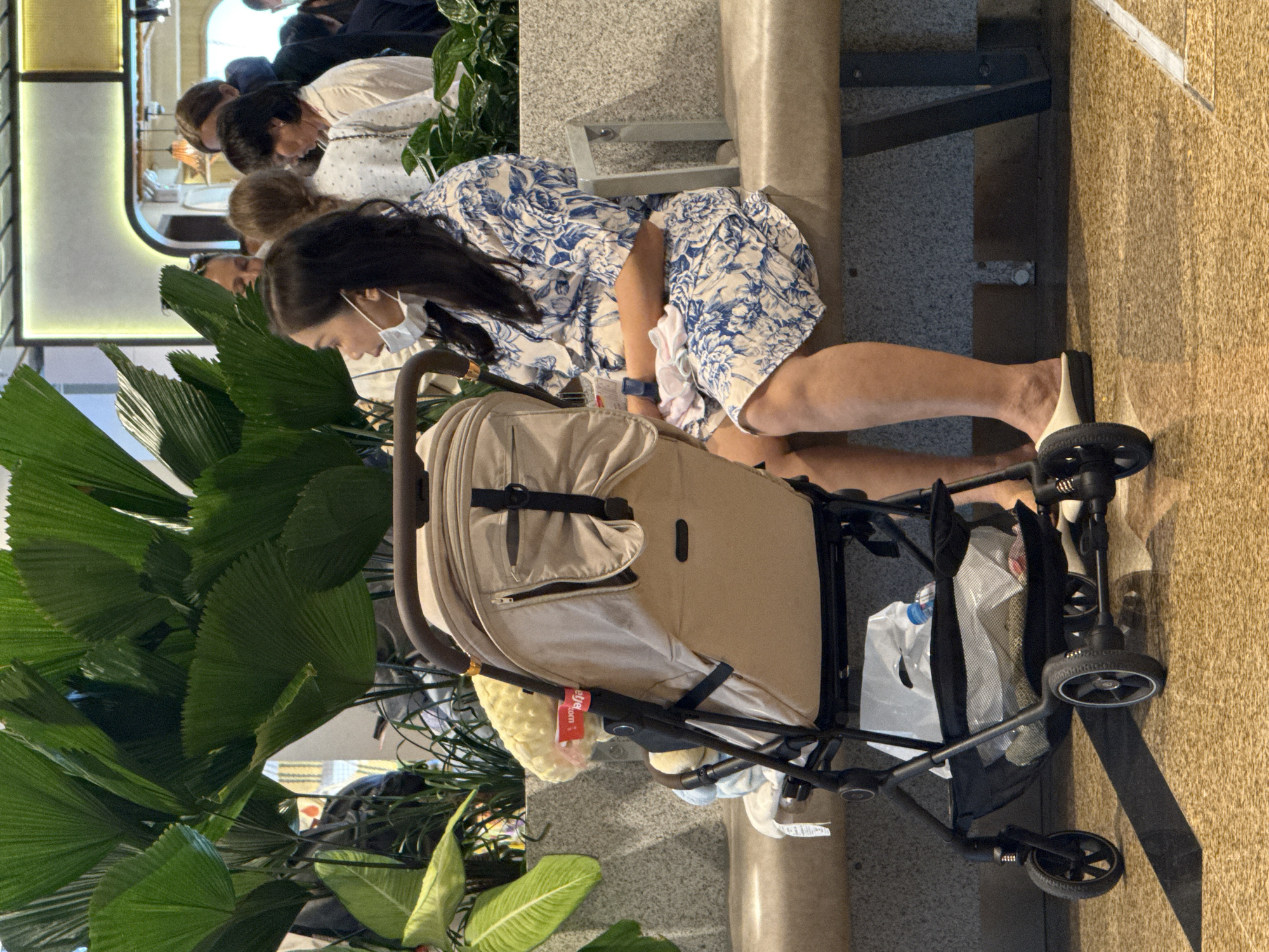 The image shows a woman sitting on a bench in what appears to be an indoor public space, possibly an airport or waiting area. She is wearing a blue and white floral dress and a face mask. A stroller is parked next to her, with a yellow blanket and some other items inside. In the background, several other people are visible, and large potted plants with fan-shaped leaves are present. The flooring is tiled. The scene is well-lit, suggesting an indoor environment. The location is identified as Bắc Cam Ranh, Vietnam.