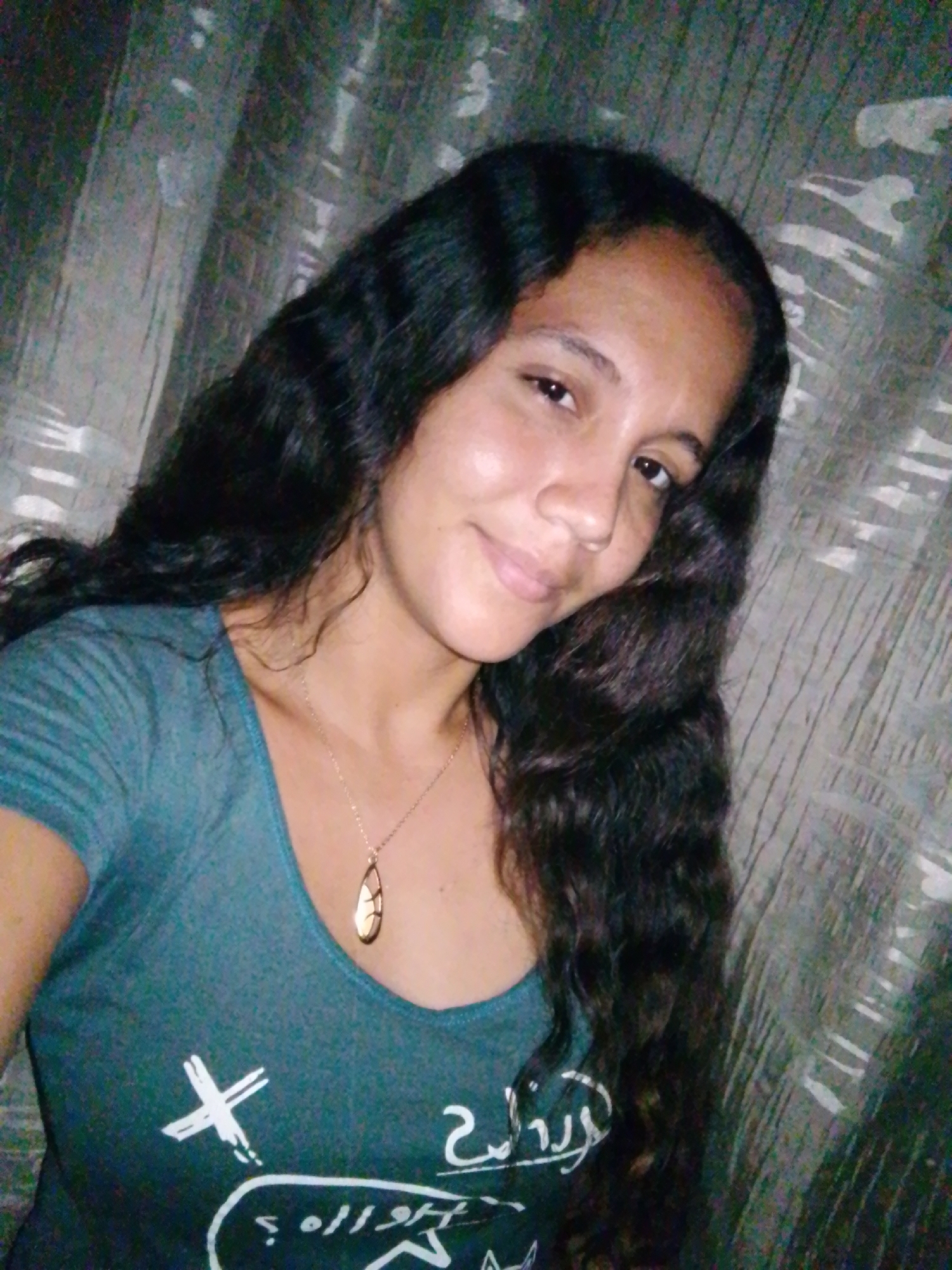 A young woman with dark, wavy hair is posing for a selfie. She is wearing a teal-colored t-shirt with a white graphic design on it and a gold necklace with a teardrop-shaped pendant. The background appears to be a wall with a textured or patterned covering. This image does not have sufficient metadata to determine that it was taken in Juan Griego, Venezuela.