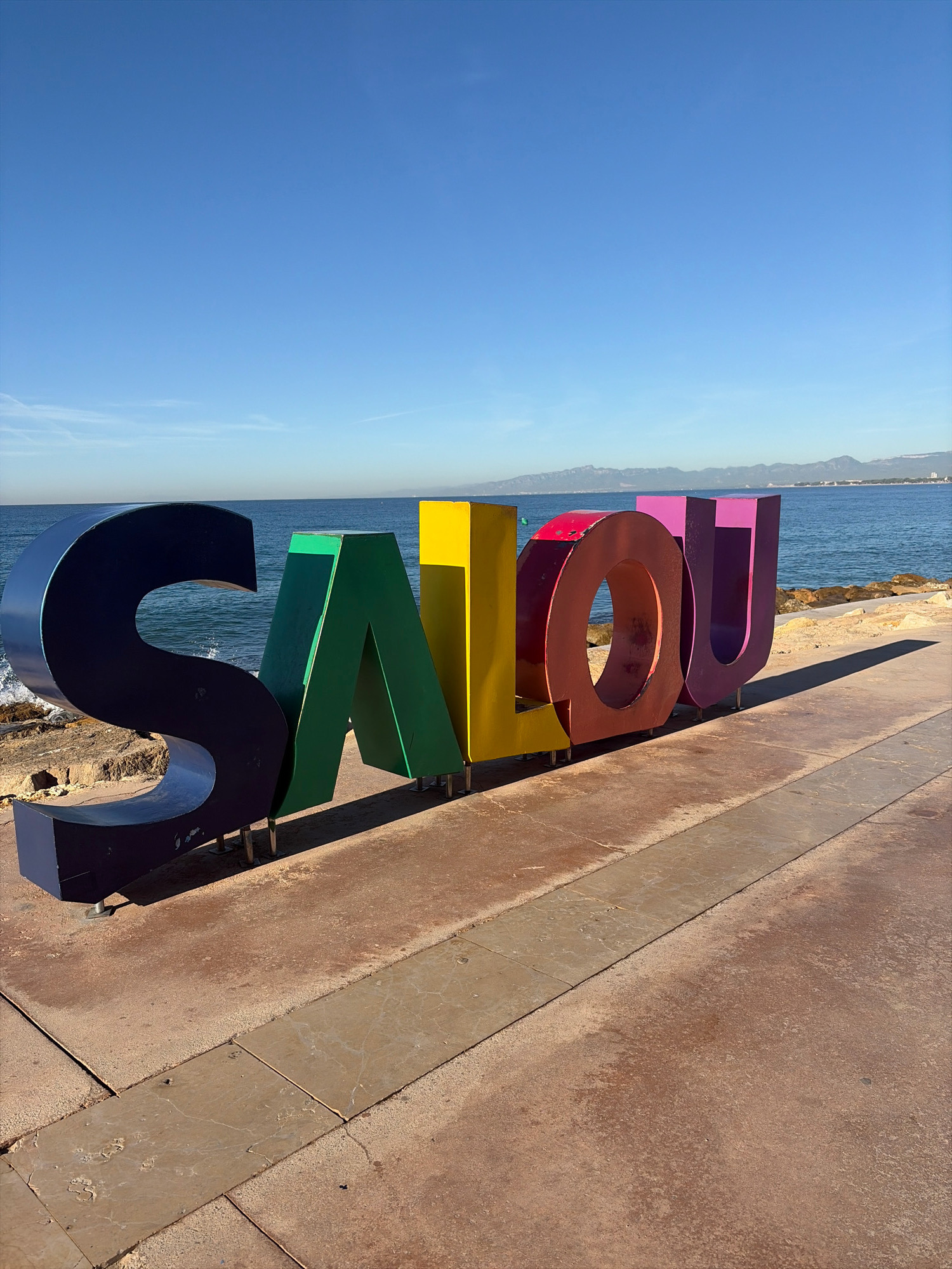 In Salou, Spain, large, colorful, three-dimensional letters spelling out "SALOU" are displayed along the waterfront promenade. The letters are constructed from metal and painted in various bright colors: dark blue for "S", green for "A", yellow for "L", red for "O", and purple for "U". They are positioned on a paved walkway that runs alongside the Mediterranean Sea. The sea is visible in the background, with a distant coastline and mountains under a clear blue sky. The sun casts long shadows from the letters onto the walkway, indicating it is likely late afternoon. The foreground shows the walkway itself, with a textured, reddish-brown surface and sections of lighter-colored, smooth paving stones. There are no people visible in the image.