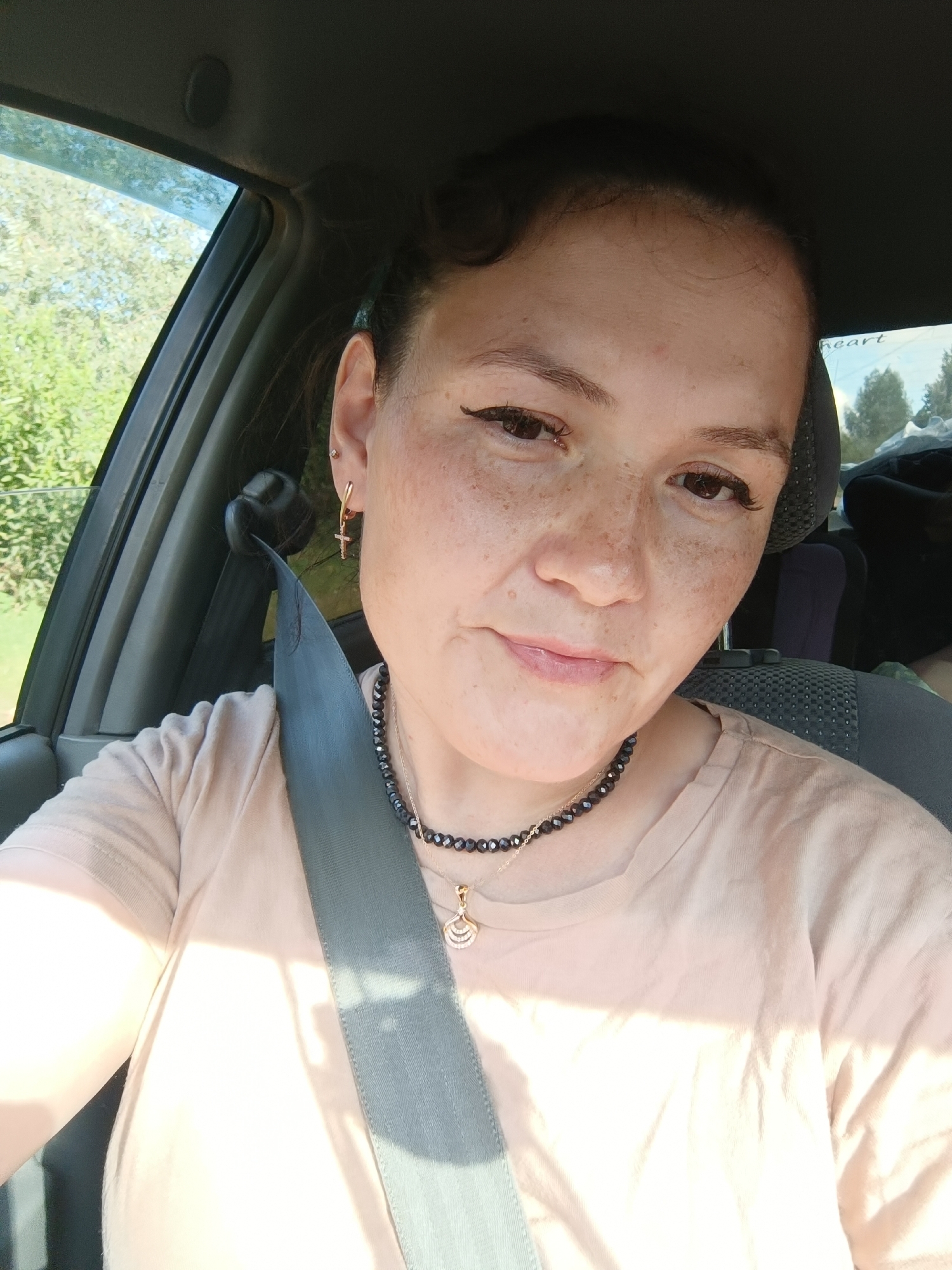 A medium shot captures a woman in the driver's seat of a car, with a seatbelt across her chest. She is looking directly at the camera with a slight smile. She has brown eyes, light brown hair pulled back, and a face dotted with freckles. She is wearing a light tan t-shirt, a black beaded necklace with a gold pendant, and gold cross earrings. The background shows trees and foliage through the car window, suggesting an outdoor setting. The image was taken in Chumna, Russia.