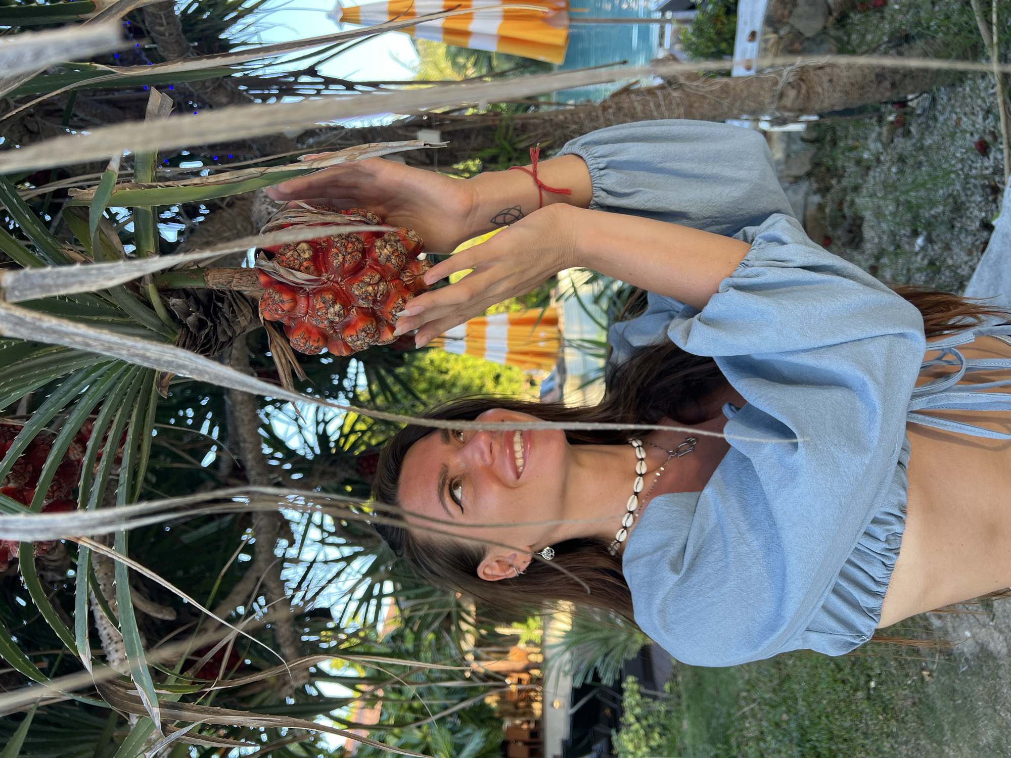 A woman with long brown hair, wearing a light blue crop top with cinched sleeves and a shell necklace, stands outdoors, holding a red, segmented fruit from a pandanus tree. She is looking up and to her right with a smile. In the background, a swimming pool and orange and white striped umbrellas are visible, along with lush greenery. The scene suggests a tropical resort or beach location in Ko Pha Ngan, Thailand.
