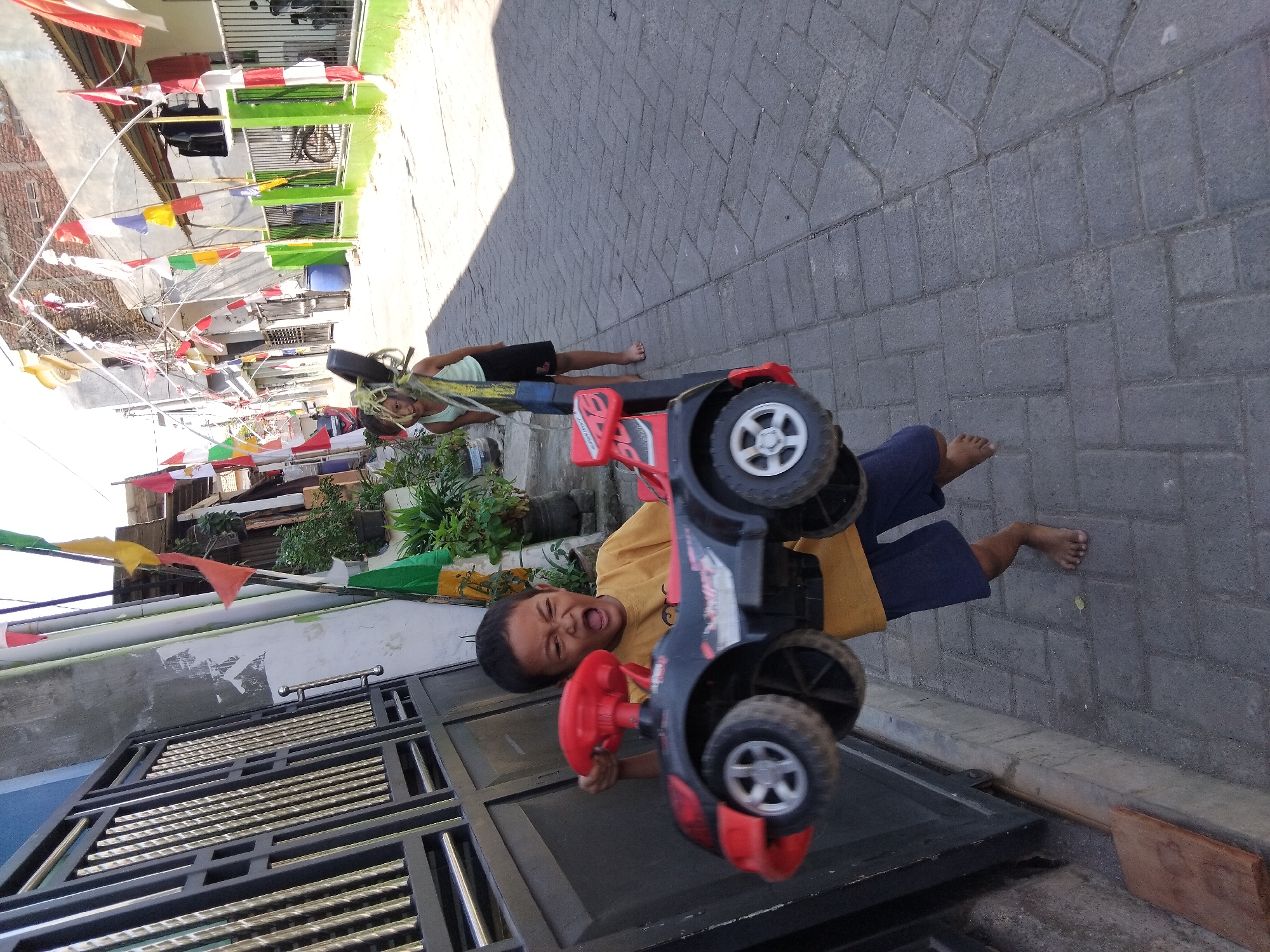 In Kepuh Kiriman, Indonesia, a young boy in a yellow t-shirt and blue shorts is enthusiastically holding a toy car, his mouth open in an excited expression. The toy car is black and red with prominent wheels. To the right of the boy, a girl wearing a light green tank top and dark shorts stands, her hair partially obscured by a toy. The scene unfolds on a paved walkway lined with houses and decorated with numerous Indonesian flags, suggesting a festive or patriotic occasion. Lush green plants in pots are visible along the path, and a dark metal gate is present on the left side of the image. The environment appears to be a residential street with brick paving and buildings in the background.