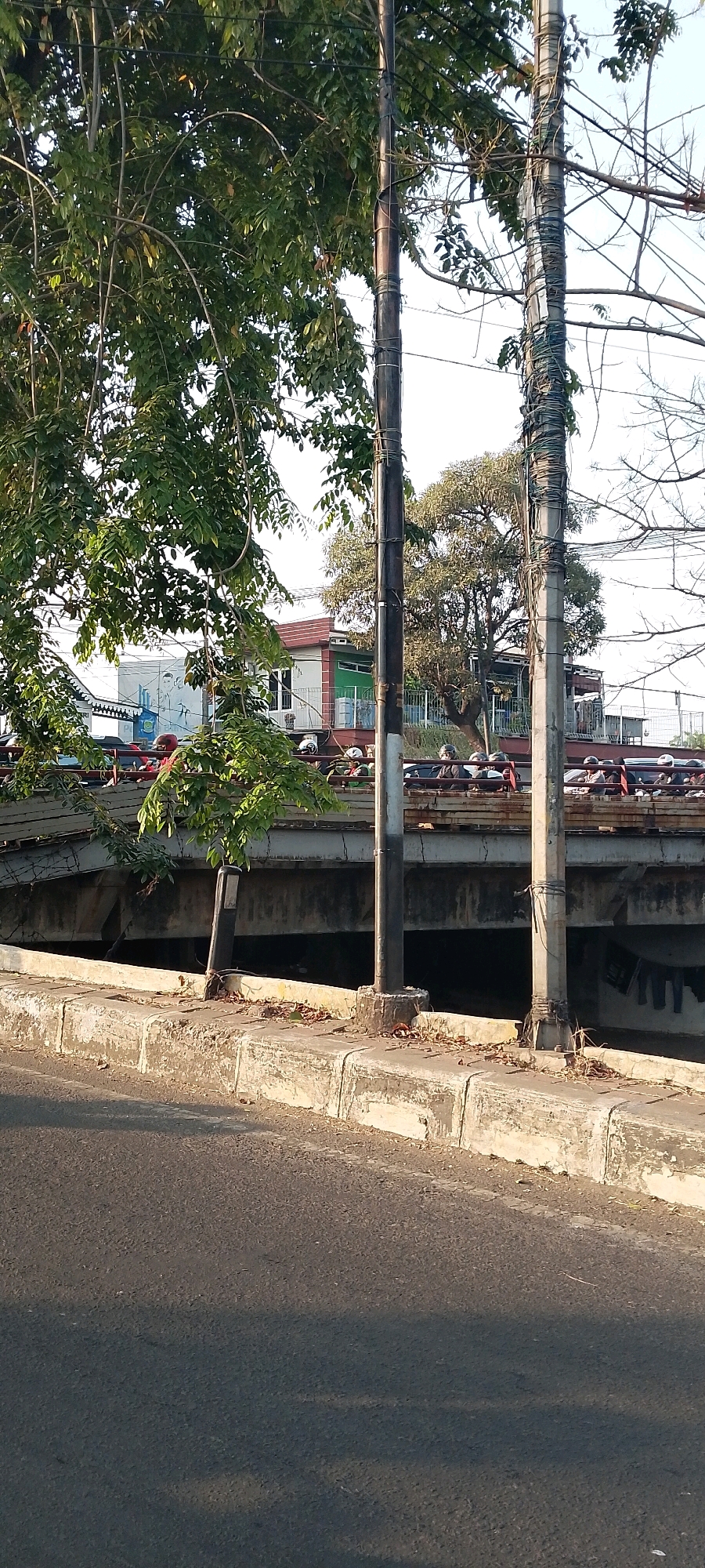 The image depicts a view of a road with a concrete curb and sidewalk in the foreground. Beyond the curb, there is a bridge structure, appearing to be an overpass. The bridge has a solid concrete underside and a railing on top. On the bridge, there are many motorcycles and their riders, suggesting moderate traffic. The scene is framed by trees with green foliage on the left and right sides, and utility poles with numerous wires are visible. In the background, behind the bridge, there is a building with a red roof and a white facade, as well as more trees. The overall impression is an urban or semi-urban environment in Bekasi, Indonesia, during daylight hours, likely late afternoon or early morning given the low angle of the sun casting shadows.
