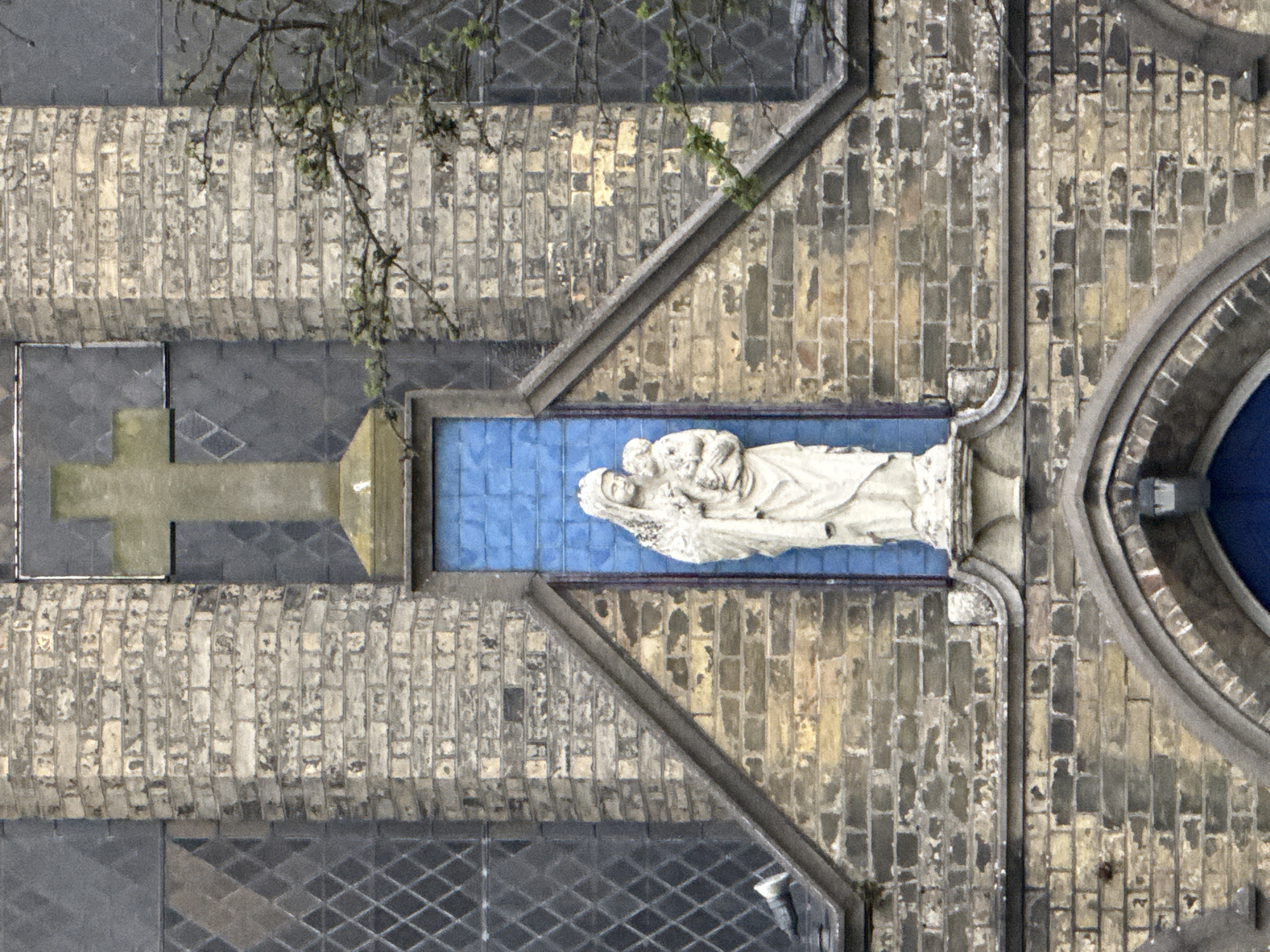 The image depicts an overhead view of a brick church structure. A stone cross is visible extending from the building's facade. A rectangular blue panel is inset into the building, showcasing a white stone sculpture of a woman holding a child. The surrounding architecture includes patterned windows and rounded architectural elements. A bare tree branch extends into the top left corner of the frame. Given the architectural style, the location is likely in the city of Great Yarmouth, United Kingdom.