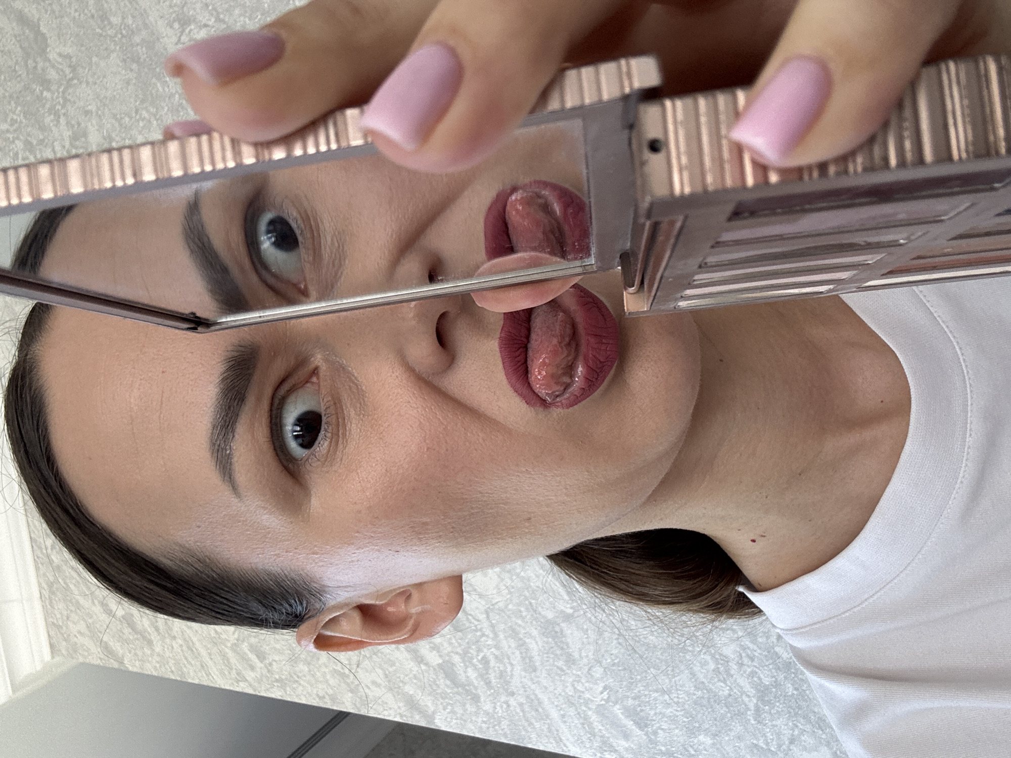 An image, captured from a slightly tilted, overhead perspective, depicts a person, likely female, engaged in self-observation using a compact mirror. The subject's face is prominent, featuring dark hair, well-defined eyebrows, and dark eyes. They are wearing a simple white t-shirt.
The person holds a small, rectangular mirror, which appears to be part of a makeup palette due to its rose gold or copper-toned, ridged casing. Their fingers, adorned with neatly manicured pink, oval-shaped nails, grasp the top edge of the palette.
A clear reflection is visible within the mirror. The reflection shows one of the person's eyes looking intently, and the top portion of their tongue, which is curled or folded and protrudes from their mouth. In the main view of the person's face, their tongue is also visible, sticking out, appearing textured, and a deep reddish-purple hue, possibly from lipstick or its natural color. This action suggests a playful or informal moment, perhaps during a routine like applying makeup or examining oral health.
The background shows a light-colored, subtly textured wall, possibly tiled, indicating an indoor setting such as a bathroom or personal space. There are no specific location cues within the image itself, other than the general interior environment. No visible text is present in the media file. The context provided indicates the location is Sochi, Russia.