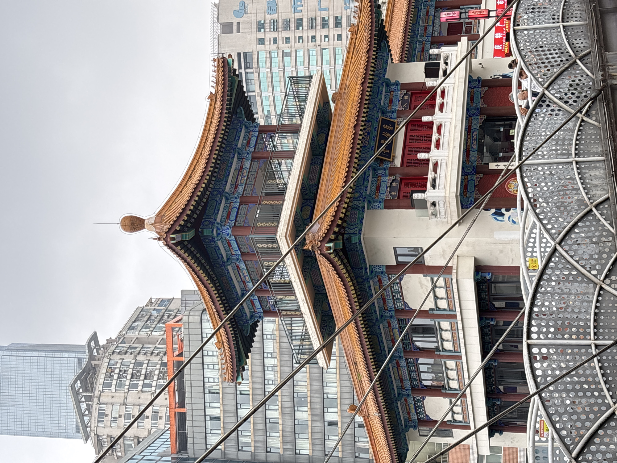 The photograph depicts a traditional Chinese pagoda-style building juxtaposed against modern skyscrapers in the Yuzhong District of China. The pagoda features ornate, curved roofs with golden tiling and intricate blue, red, and white decorative elements. A metal structure with a grid-like pattern is visible in the foreground, partially obscuring the pagoda. Behind the pagoda, modern buildings rise with glass and concrete facades, indicating a mix of historical and contemporary architecture in the urban landscape. Overhead wires are present throughout the scene. Several people stand along a landing on the pagoda.