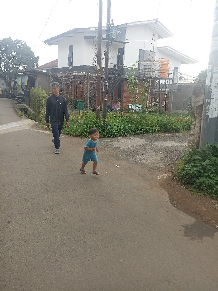 The image shows a street scene in Jatinangor, Indonesia (latitude: -6.937, longitude: 107.779). A young boy, wearing blue shorts and a blue shirt, is running in the center of the street. Behind him, an adult male is walking in the same direction. The adult male is wearing a dark colored shirt, dark pants, and sneakers. To the right of the scene is a concrete wall and a green, grassy area. To the left, there is a house with a white facade and brown roof. The house has a small, metal railing. It has a small, metal balcony. The street is paved with dark asphalt. The sidewalk is a light grey color. The weather is overcast and cloudy. The sky is a light grey. The air is clear. The houses are set back from the road. The boy is running away from the man.