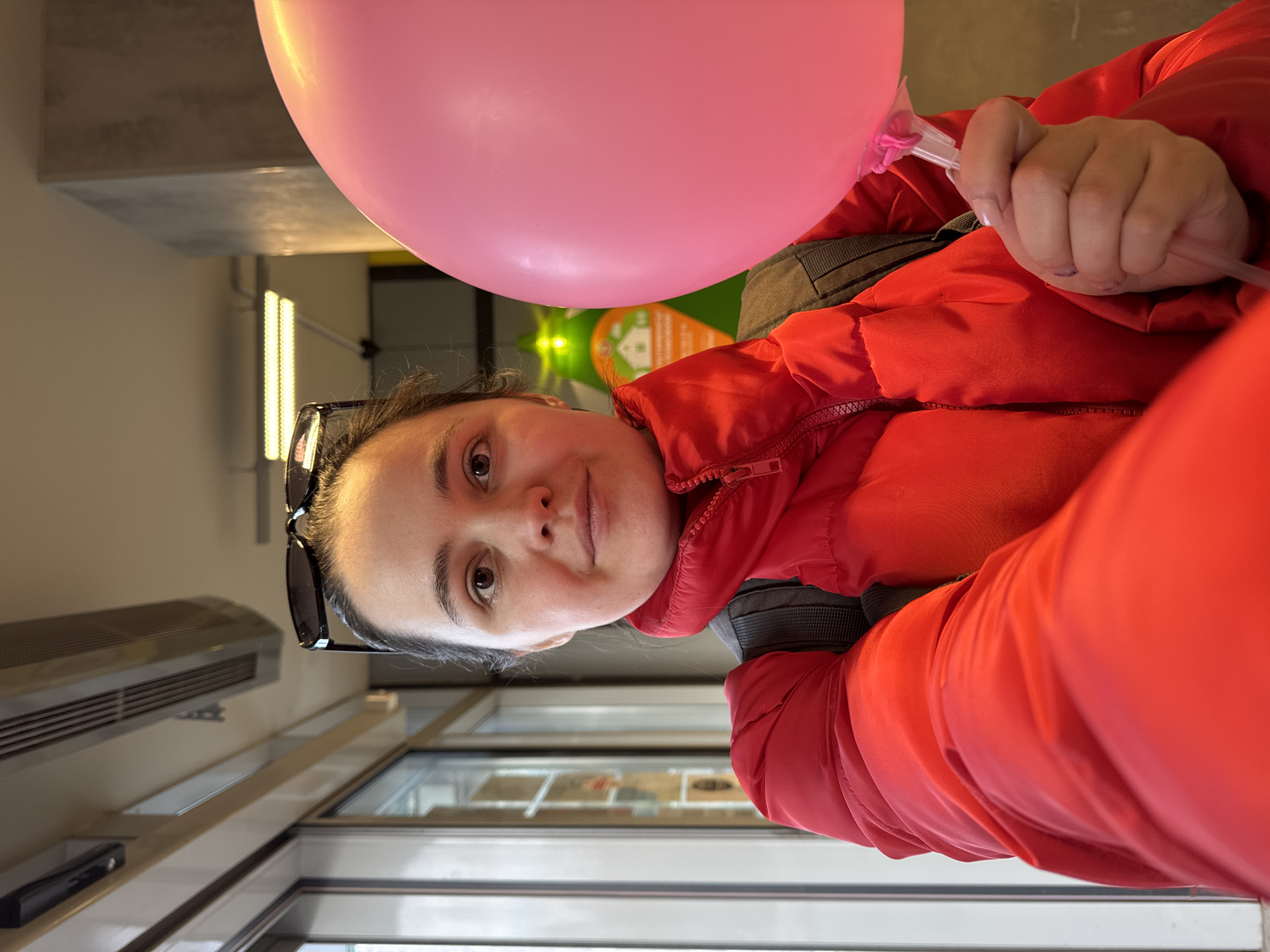 A woman wearing a red puffer jacket and sunglasses on her head is holding a pink balloon. She is standing inside a building with glass doors and windows. The location is Izhevsk, Russia. There is an air conditioning unit and fluorescent lights visible in the background. A green sign is also present in the background.