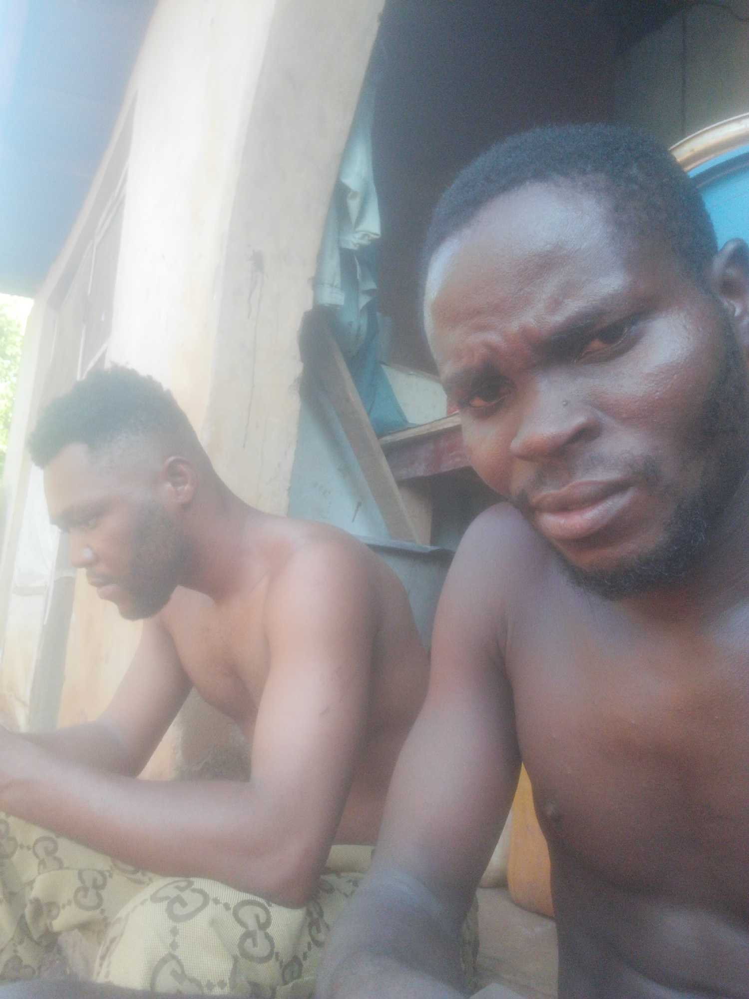 The image features two shirtless men of dark complexion, likely outdoors or in an open-air setting, during daytime. The lighting is bright and natural.
In the foreground, a man is positioned close to the camera, looking directly into it with a somewhat intense or serious expression. He has short dark hair and a beard. His right shoulder and arm are visible.
In the background, another man is seated, seen in profile looking downwards and to his left. He also has short dark hair and a beard. His torso is bare, and his lower body is covered by a fabric that displays a repeating pattern of interlocking shapes resembling the letters "G" or "GG", commonly associated with the Gucci brand.
The background shows a light-colored wall or structure, possibly made of concrete or plaster, with some architectural details like an archway. To the right of the background man, there are some wooden structures and draped blue fabric. The overall setting appears somewhat rustic or simple. No explicit text is clearly legible other than the pattern on the fabric.