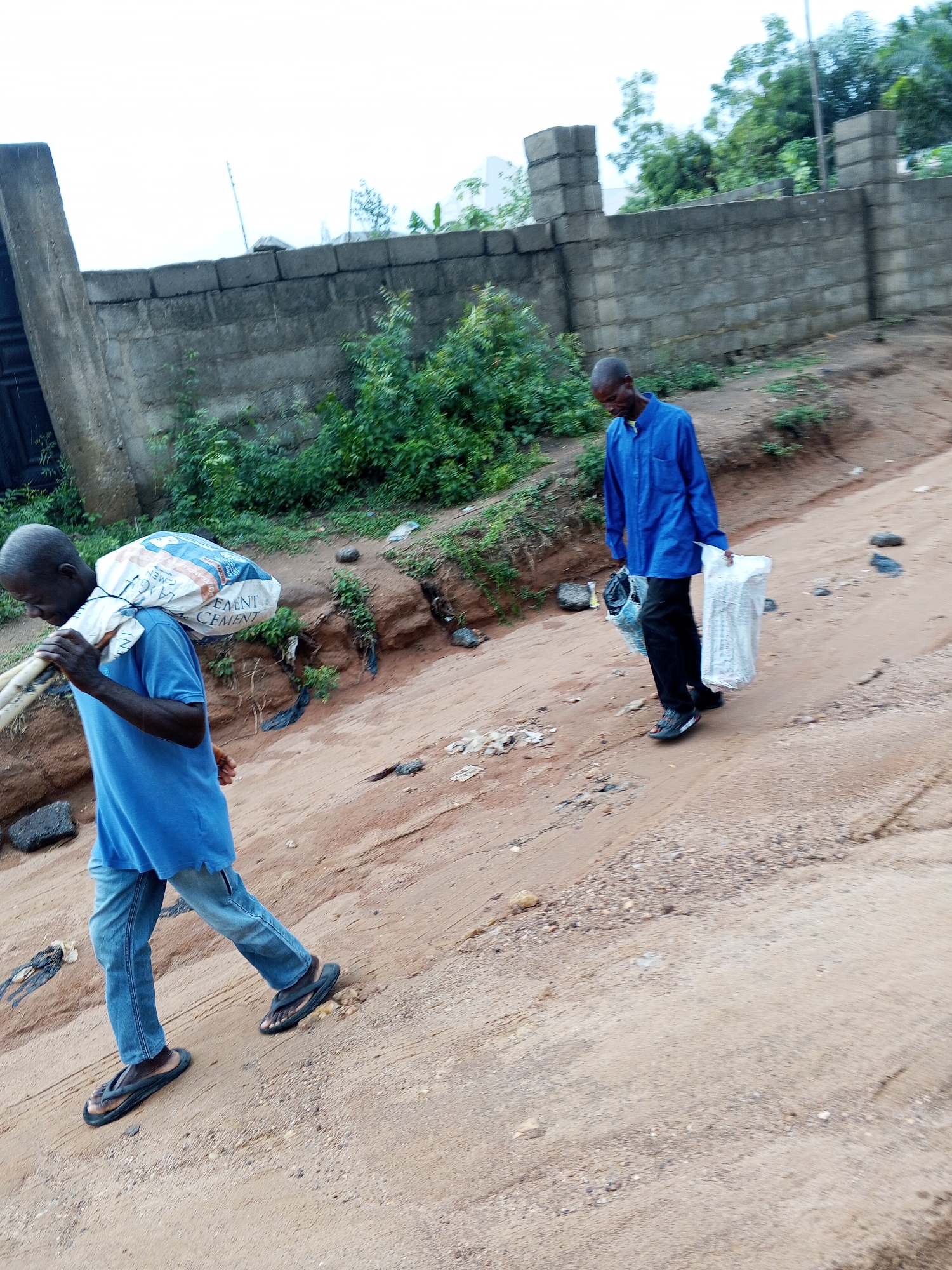 Two men are walking along a dirt road. The man on the left is carrying a bag of cement on his shoulder. The man on the right is carrying plastic bags in his hands. The road appears to be slightly elevated. In the background is a concrete wall with vegetation. This is in the city of Zango-ndaji, Nigeria.