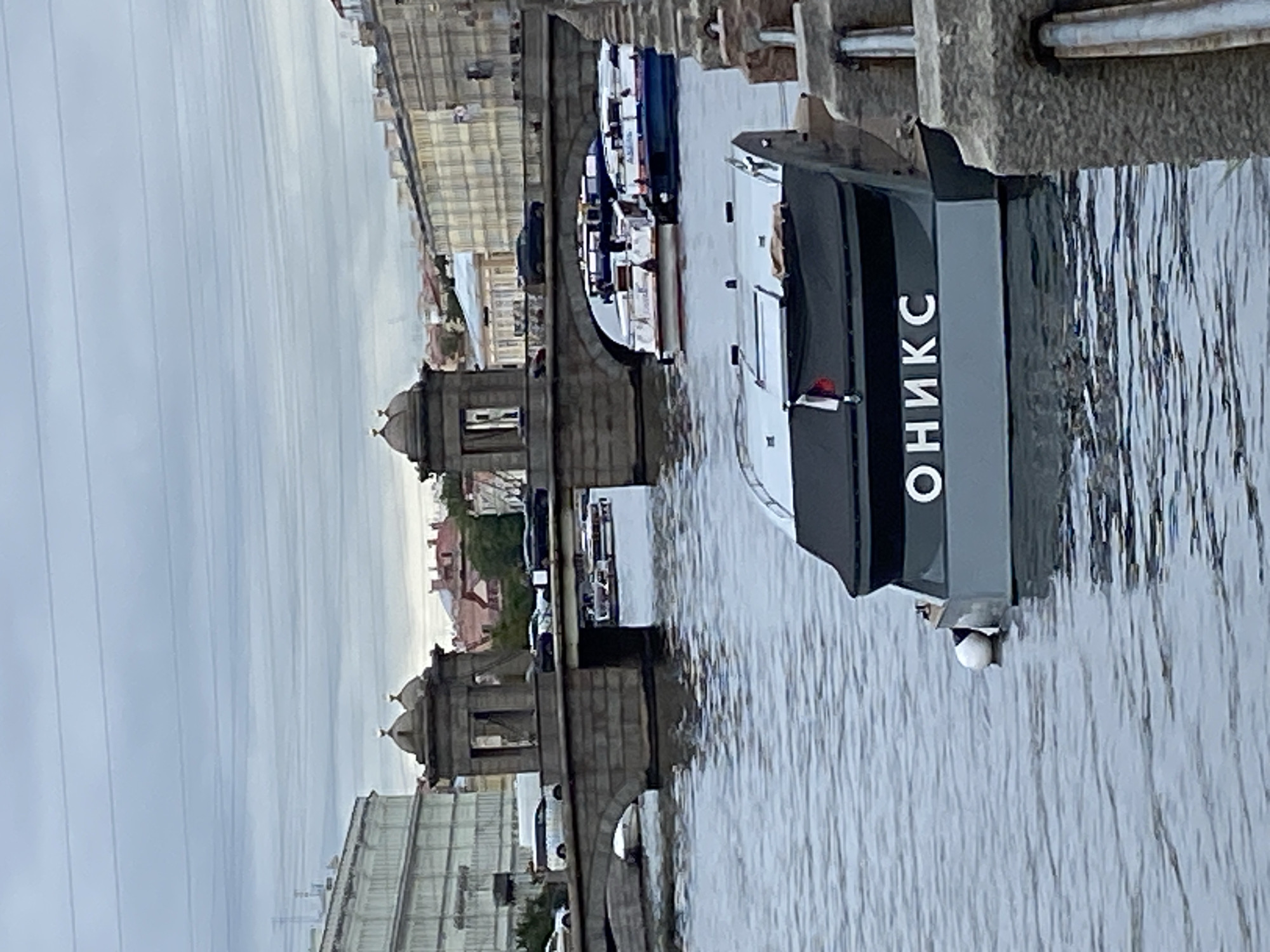 The image captures a view of a canal in Saint Petersburg, Russia. A gray barge with the Cyrillic word "ОНИКС" (ONYX) in white letters emblazoned on its side is moored in the foreground. The barge has a black canopy and a small Russian flag. In the mid-ground, a stone bridge spans the canal, with vehicles including cars and vans visible on its roadway. Two passenger boats are docked beneath the bridge arches. The background features buildings with classical architecture, typical of Saint Petersburg, and a cloudy sky. The canal is calm, reflecting the overcast conditions and the surrounding structures. The overall scene depicts a tranquil urban waterway with visible boat traffic.