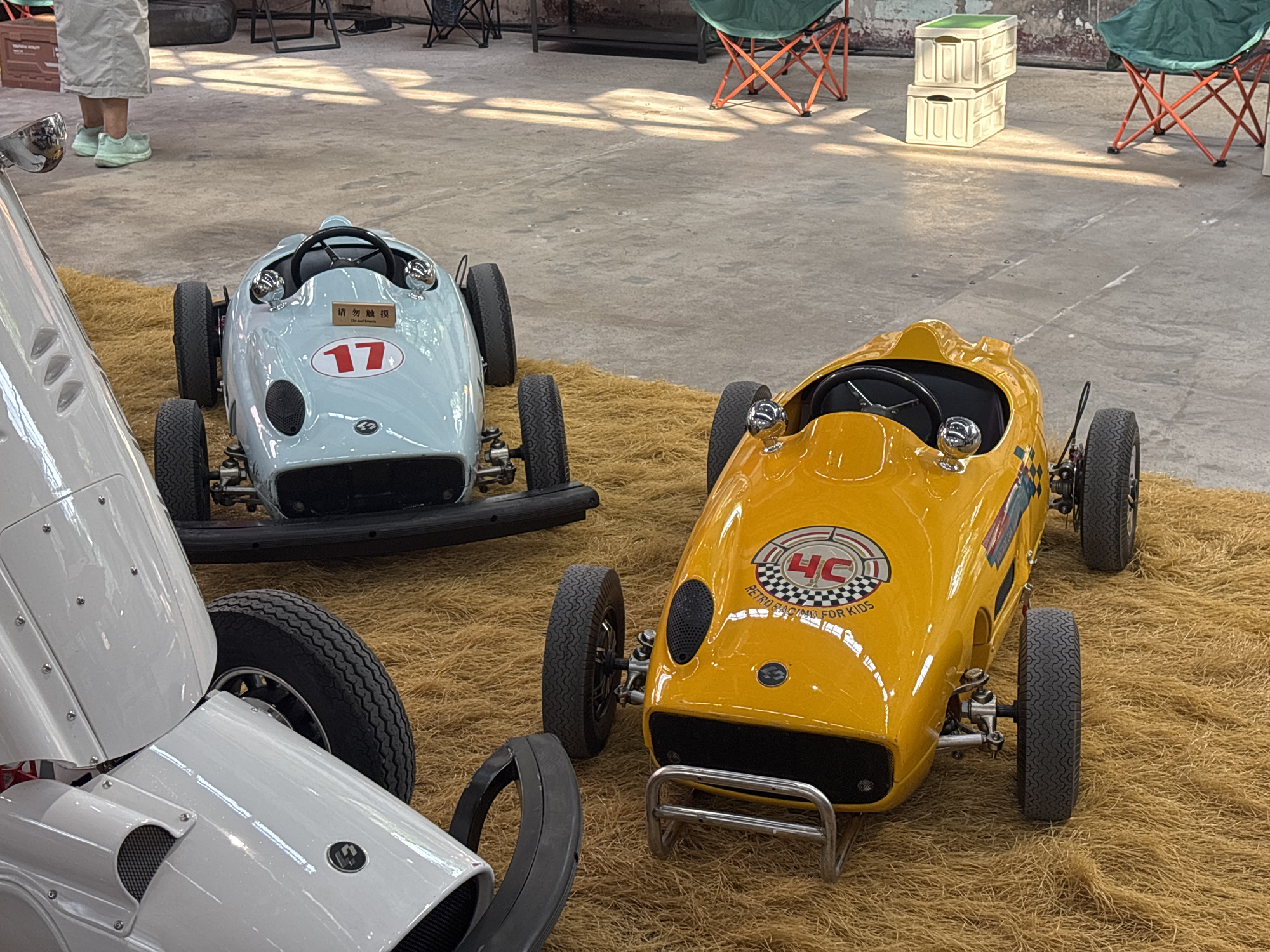 The image depicts a scene with several pedal cars, likely intended for children, placed on a tan, fibrous matting. Two of the pedal cars are prominently visible: one light blue and one bright yellow. The blue car, positioned on the left, features a visible number "17" and has a sign with Chinese characters and the English phrase "Do Not Touch." The yellow car, on the right, displays a circular logo with "HC" and checkered patterns, along with text that appears to read "RETRO RACING FOR KIDS." A portion of a white pedal car is visible in the foreground on the left. The background shows a concrete floor with scattered shadows, suggesting an indoor or partially covered outdoor setting. In the far background, there are foldable orange chairs with green seats, and stacked white boxes. A person wearing light-colored pants and mint green shoes is partially visible in the upper left corner, standing near the blue pedal car. The overall impression is that of a display or exhibition of vintage-style toy vehicles. The context of Pudong, China, suggests this might be a commercial or event space within that district.