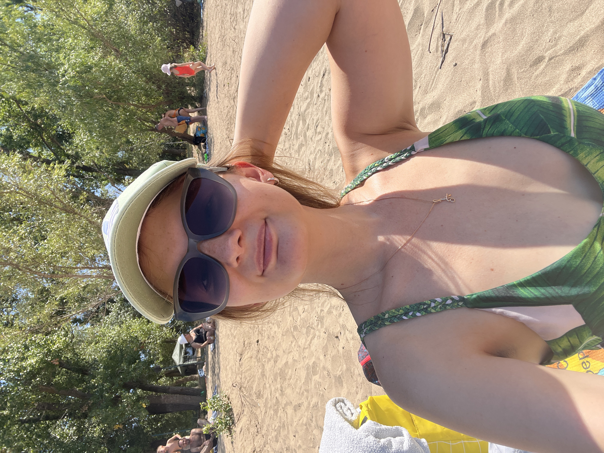 The image depicts a selfie of a young woman wearing a light green baseball cap, large sunglasses, and a green and white patterned swimsuit. She is on a sandy beach, with trees and other people visible in the background. To her left, a yellow bag and a white towel are placed on the sand. In the background, several individuals are seen relaxing on the beach, some sitting and others standing. The environment appears to be a sunny day in a natural setting, likely a beach by a body of water. The location is identified as the city of Kilyakovka, Russia.