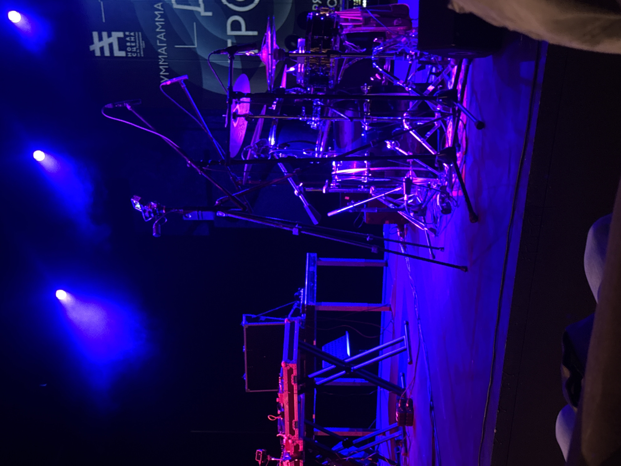 On a dimly lit stage, bathed in vibrant blue and pink spotlights, a drum set, microphones on stands, and a keyboard setup are arranged. The background features a large banner with Cyrillic lettering and abstract graphics, suggesting a performance or event space. A portion of the audience, blurred and out of focus, is visible in the foreground. The overall scene suggests a live music performance or a rehearsal in an indoor venue, possibly a concert hall or club in Saint Petersburg, Russia, given the Cyrillic text and the typical setup for live music events in the city.