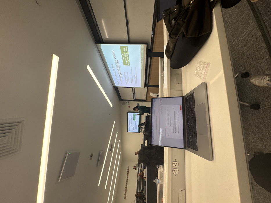 This image depicts a classroom or lecture hall scene at Babson College in Wellesley, Massachusetts, United States. The perspective is from a student's desk, looking towards the front of the room.
In the immediate foreground, a silver laptop is open on a white desk, displaying an interface titled "Application Question 1 of 5." To the right of the laptop, a small, clear plastic zip-lock baggie and a black backpack rest on the desk. Two electrical outlets are integrated into the desk's front panel. Below the desk, the feet of the person taking the photograph are visible, wearing light-colored sneakers.
In the midground, multiple students are seated in tiered rows of white desks. The heads and upper torsos of several individuals are visible, primarily with dark hair. A plastic water bottle is present on one student's desk.
At the front of the room, a female instructor, wearing a dark green top, stands behind a podium, actively gesturing with both hands towards the students. Behind her, two projection screens are visible. The main, larger screen on the right displays a presentation slide titled "Economic Order Quantity (EOQ) Model," detailing "Trade-offs: Variable vs. Fixed Costs" and "Risk Prone vs Risk Averse," with "BABSON COLLEGE" and "WWW.BABSON.EDU" at the bottom. A smaller screen to the left also displays presentation content, including a table or chart.
The classroom features a modern aesthetic with white walls, extensive linear fluorescent lighting integrated into the ceiling, and grey carpeted flooring. Ceiling vents are also visible.