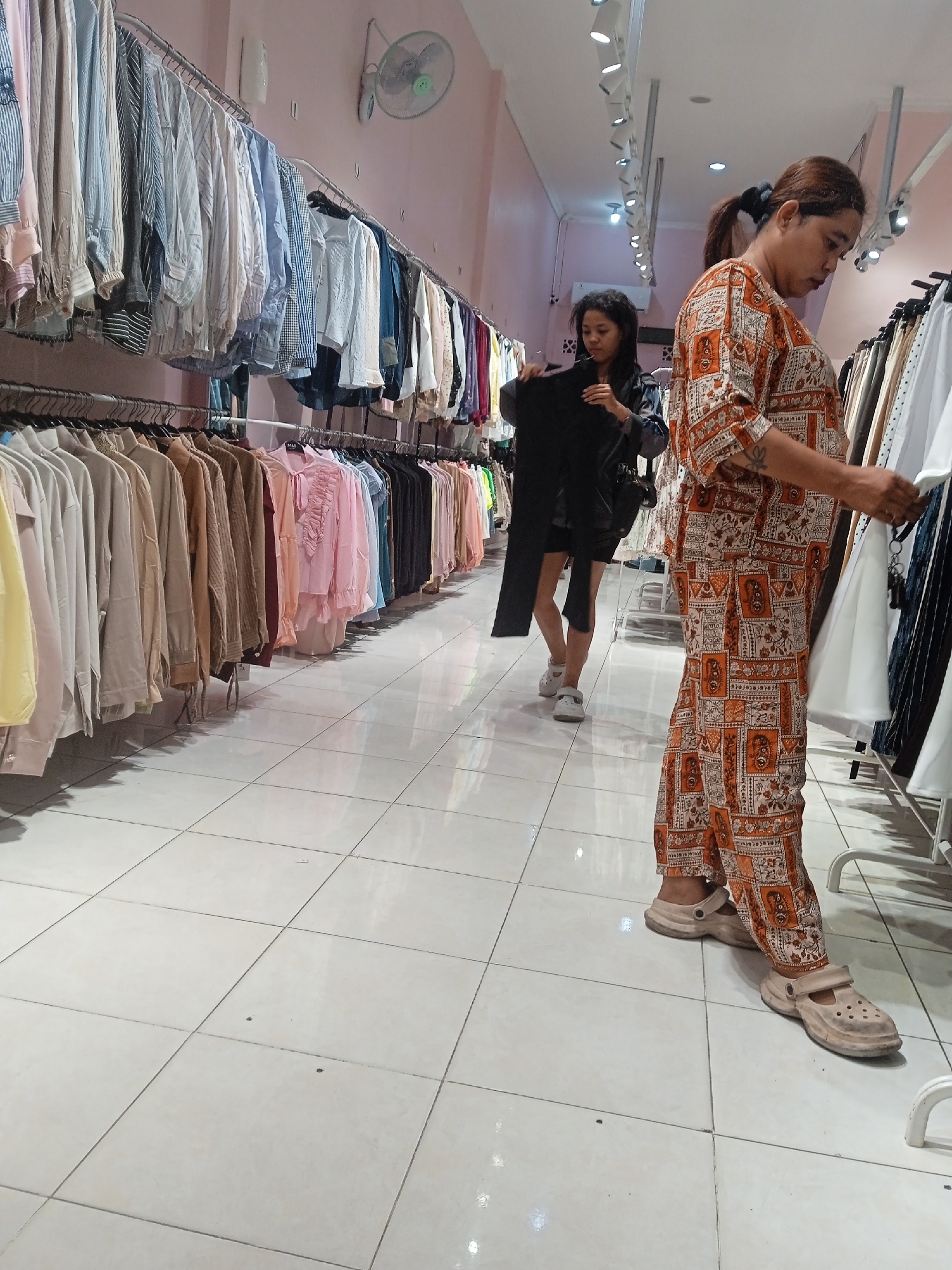 This image captures a retail scene within a clothing store in Semarang, Indonesia, where two individuals are browsing through racks of apparel. A woman, wearing a printed orange and white outfit and beige clogs, is standing and examining a white garment. Behind her, another woman, dressed in black, is holding up a black garment. The store environment is characterized by rows of clothing on hangers, illuminated by overhead lights. The floor is tiled, and a fan is mounted on the pink wall.