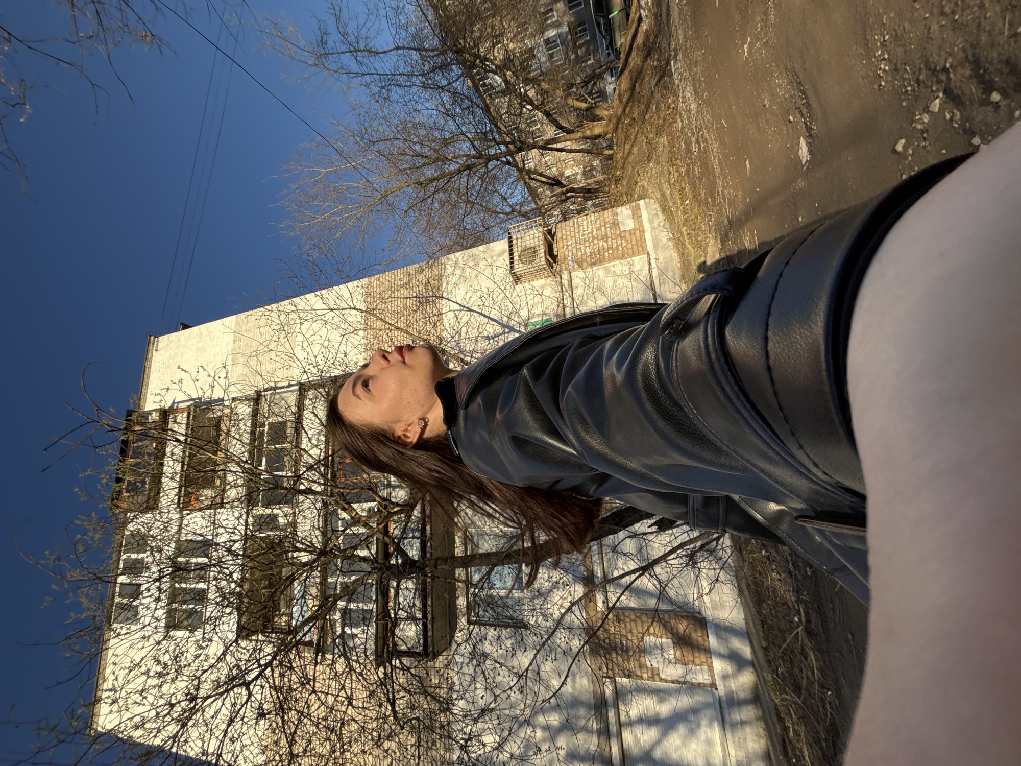 The photograph is an outdoor shot taken in Perm, Russia. The focus is on a woman with long brown hair, wearing a black leather jacket. She is positioned in the foreground, facing upwards and towards the left side of the frame. Behind her, there is a multi-story building with white walls and numerous windows, typical of urban residential architecture. Leafless trees are situated in front of the building, casting shadows on its facade. A clear blue sky is visible above the building, while a paved road or sidewalk is present on the right side of the image. The lighting suggests it is daytime, with the sun illuminating the building and the woman.