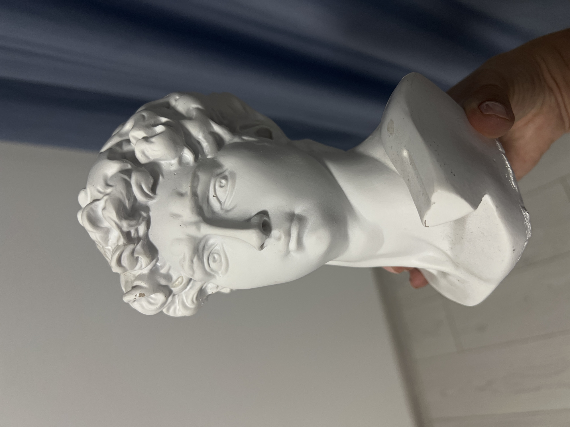 A bust of Michelangelo's David, made of white plaster, is held by a hand in the foreground. The bust is facing slightly to the left, with its head tilted down and to the left. The hair is sculpted in tight curls, and the facial expression is stern, with furrowed brows. The bust appears to be broken at the base. In the background, a grey wall and a blue curtain are visible. The floor below is light-colored wood flooring. The image appears to be taken indoors.