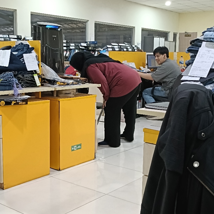 The scene is a retail store in Tugu, Indonesia, located at coordinates -6.973 latitude and 110.326 longitude. A woman wearing a red shirt and a black headscarf is bending over a yellow counter, looking at the items displayed on it. Behind her, a man wearing a grey shirt is sitting in a chair and looking at a computer screen. He has his right hand on the keyboard and his left arm on the back of the chair. Both the counter and the chair are facing the same direction. The man has brown hair and is wearing a pair of blue jeans. The woman has a large black tote bag on her shoulder. The counter is stocked with merchandise, a rack of folded blue jeans to the left and other items to the right. The background behind the woman is a wall with glass windows and a row of brown filing cabinets. There is a large black air purifier on the floor to the left of the woman. The floor is white tiled. There is a black jacket hanging on a rack in the foreground to the right of the image.
