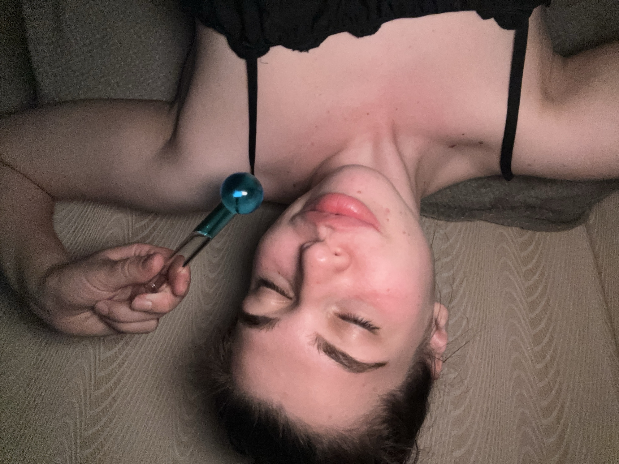 A woman is lying on a patterned surface, possibly a couch or bed. She is wearing a black top with thin straps. She is holding a blue facial ice globe in her right hand. Her eyes are closed. The area in the city of Мещёры, Russia cannot be determined.