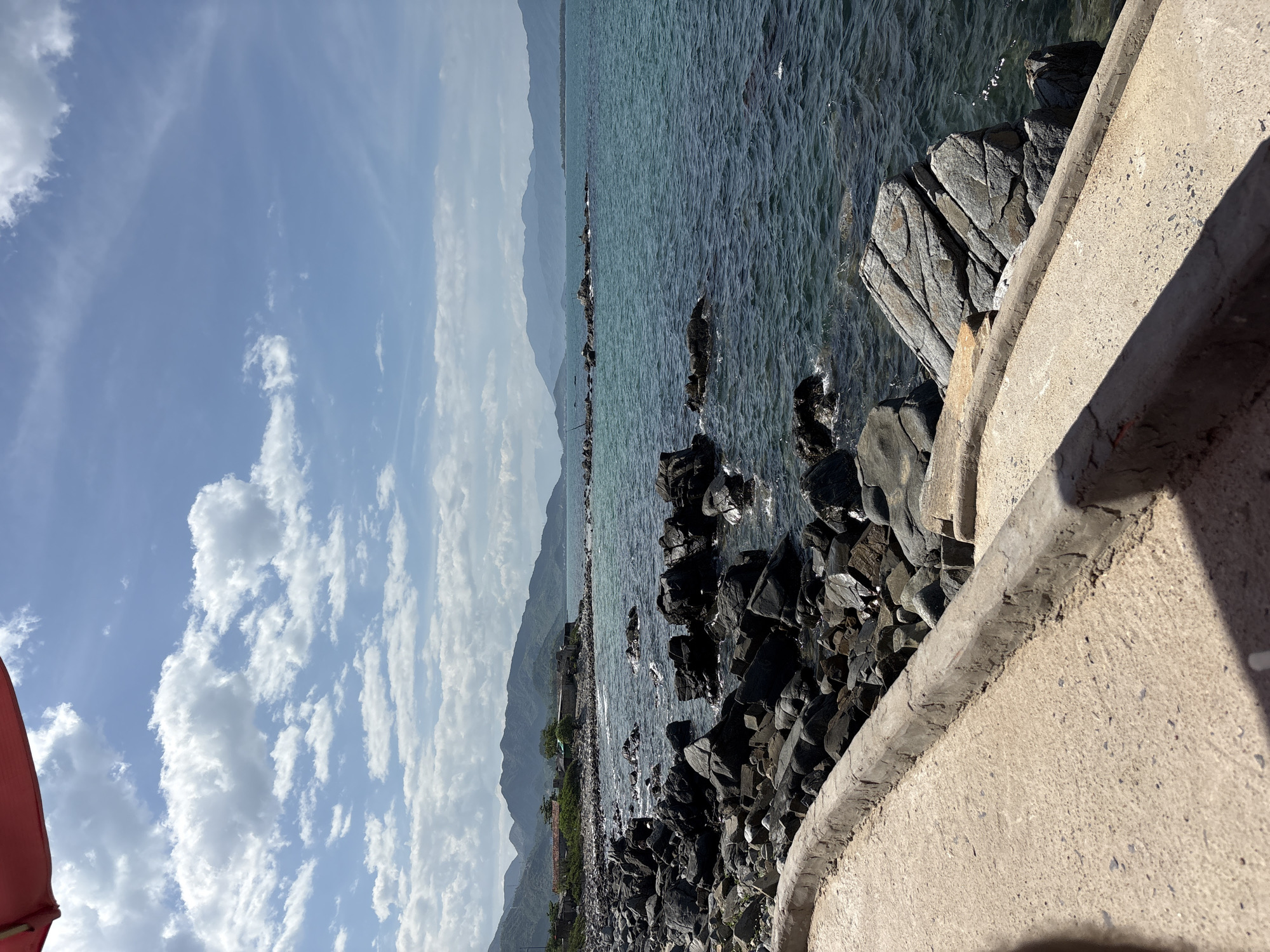 This is a vertical, eye-level shot of a rocky coastline and the ocean in Vĩnh Lương, Vietnam. The foreground features a concrete pathway with a raised, weathered edge that curves to the right. To the left of the pathway are numerous dark, irregular rocks, some of which are partially submerged in the water. The ocean is a clear turquoise color with moderate waves and some whitecaps. In the distance, a chain of mountains forms the horizon, with several small structures and trees visible along the shore. The sky is blue with scattered white, puffy clouds and a hint of a red awning in the upper left corner.