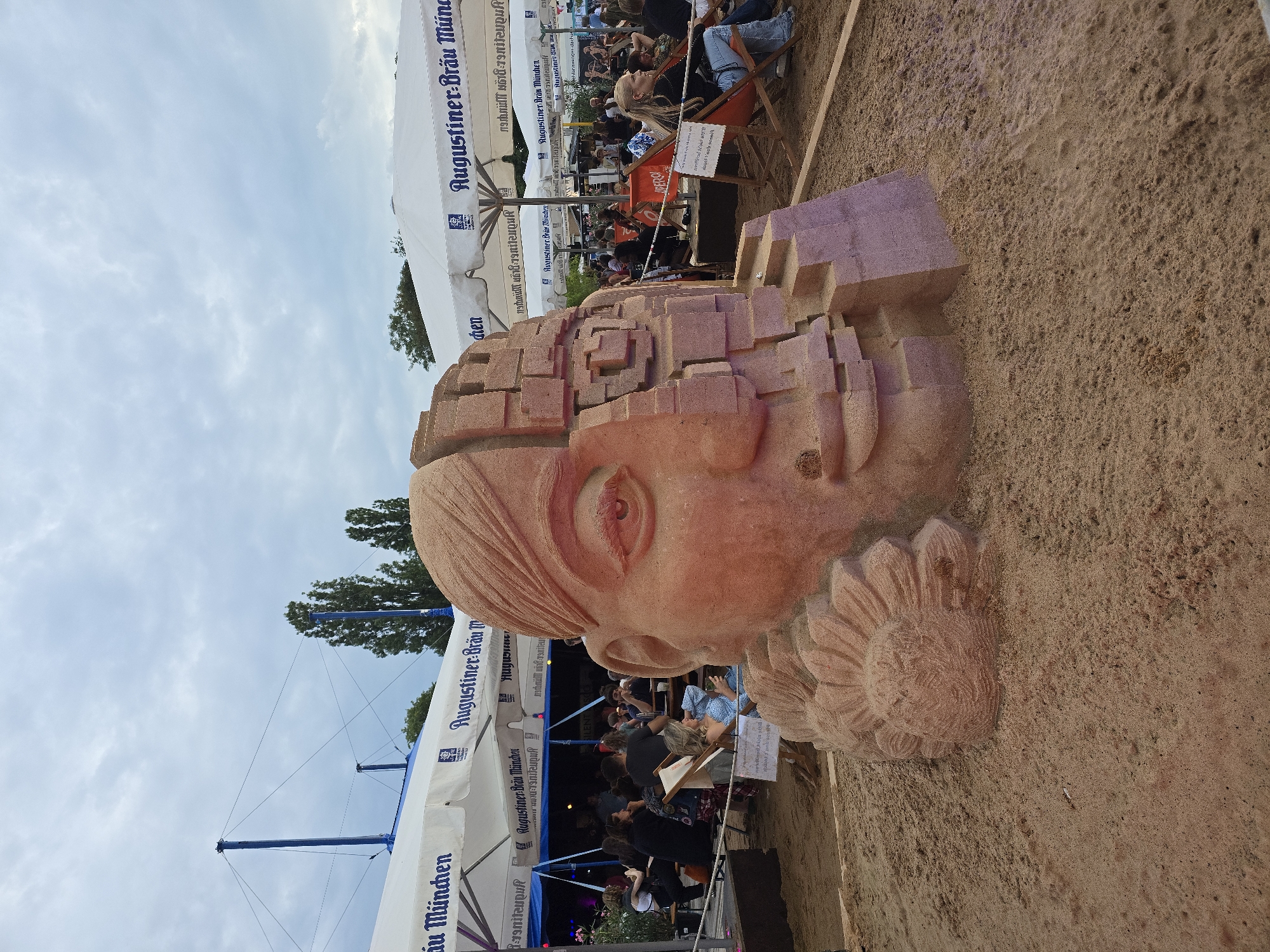 This is a vertical, close-up, outdoor photograph of a sand sculpture of a human head, with the background slightly blurred. The sculpture is made of reddish-brown sand and depicts a face with detailed features, including an eye and a sunflower-like design on the side of the head. The head is fragmented, with parts of it appearing to be made of blocks, suggesting a digital or deconstructed theme.
In the background, there are white tents with blue lettering that reads "Augustiner Bräu München," indicating a beer garden or festival setting, possibly in Munich, Germany. People are visible sitting at tables and on chairs under the tents, suggesting a daytime event. The sky is partly cloudy, suggesting mild weather. The overall impression is of an art installation at a public gathering.