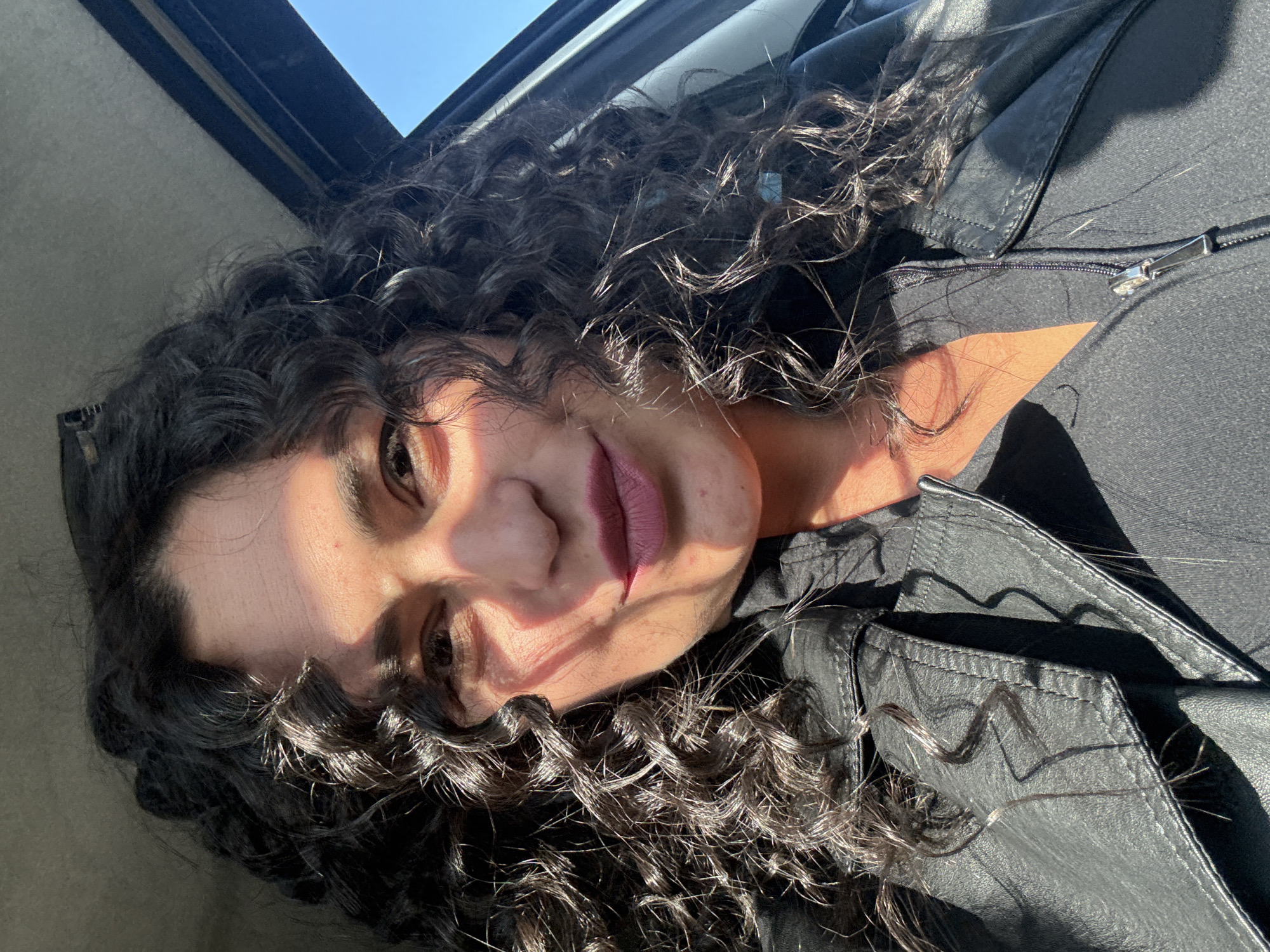 A close-up, tilted portrait shows a person with abundant dark, curly hair, illuminated by strong, direct sunlight. The lighting casts distinct shadows across the face, highlighting one eye and lips painted with a dark shade, which display a slight, relaxed smile. Individual strands of their hair catch the light, revealing texture and shine.
The person is wearing a black jacket, possibly made of leather or a similar material, with a visible zipper down the front. The background suggests an interior space, likely a car, with a partial view of a sunroof or window frame and a clear blue sky beyond. The bright conditions indicate it is daytime with sunny weather. The perspective is from above or with the subject looking upwards. No text is visible in the image. The location context for the image is Кемуль, Russia.
