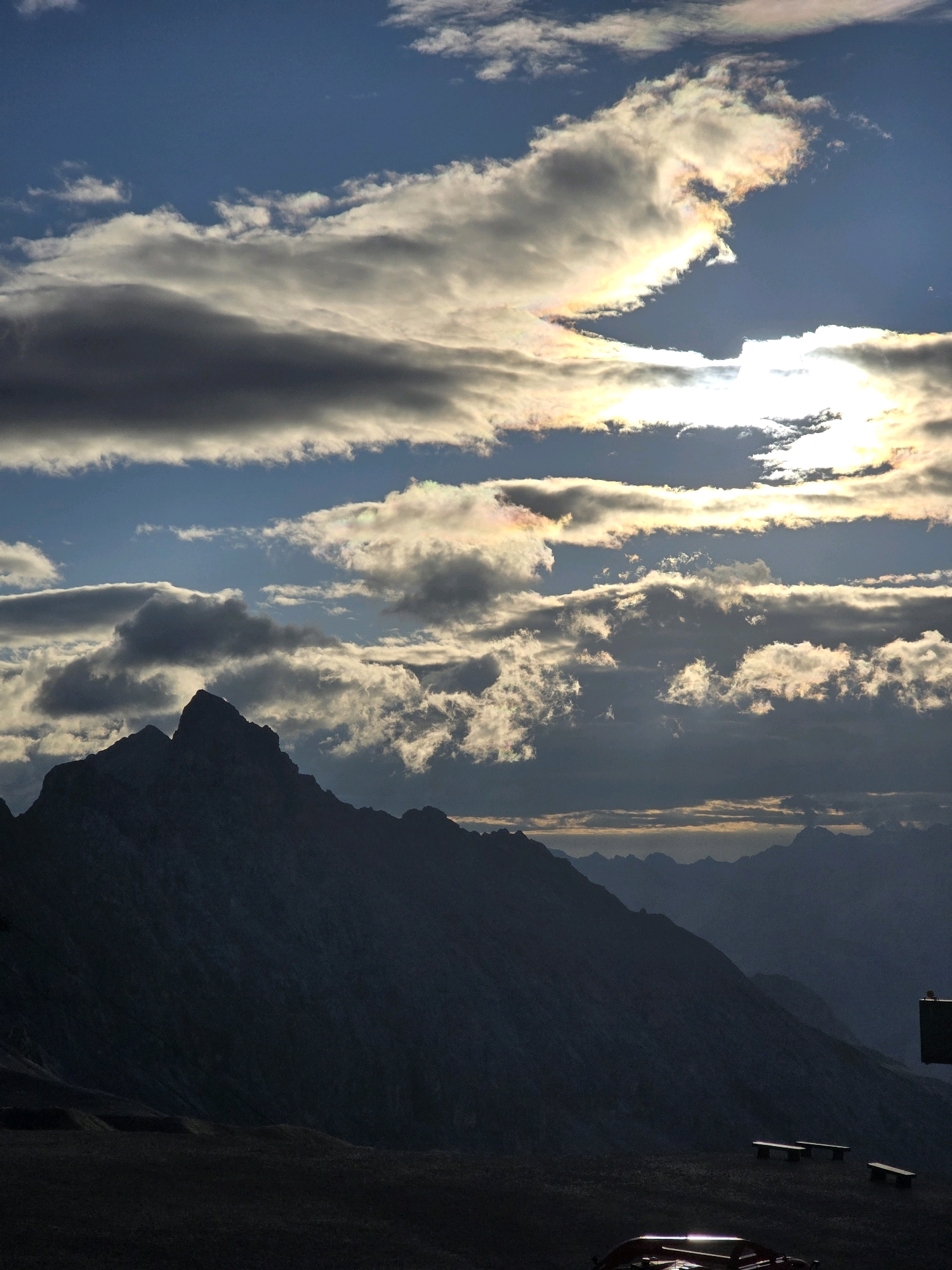 The image depicts a mountainous landscape under a cloudy sky. In the foreground, a dark silhouette of a mountain range dominates the lower portion of the frame. The sky is filled with a variety of clouds, some of which are illuminated by sunlight, creating iridescent effects. A few smaller, silhouetted mountain peaks are visible in the distance. In the bottom right corner, part of a structure, possibly a building or observation deck, is visible, along with what appear to be benches in the mid-ground. The overall impression is one of a dramatic natural scene, possibly taken at sunrise or sunset. The location is identified as Garmisch-Partenkirchen, Germany. There are no people visible in the image.
