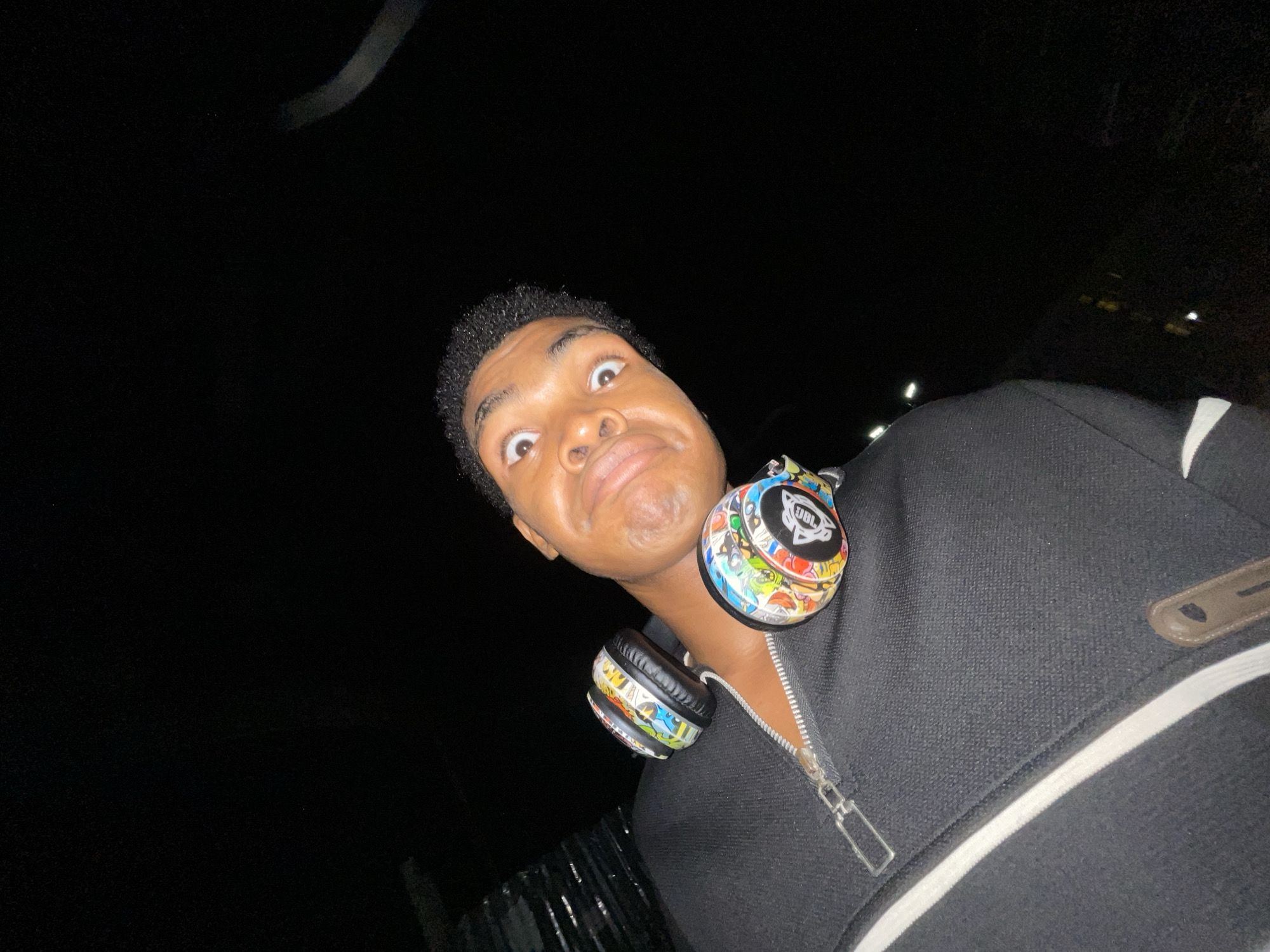 The image shows a young Black male wearing a black zip-up jacket with a white stripe and what appear to be JBL headphones decorated with stickers around his neck. The man is looking directly at the camera with wide eyes and a slightly distorted facial expression. The background is mostly dark, suggesting it is night, and the location may be Ilado, Nigeria, as requested. There is a faint light source above him.